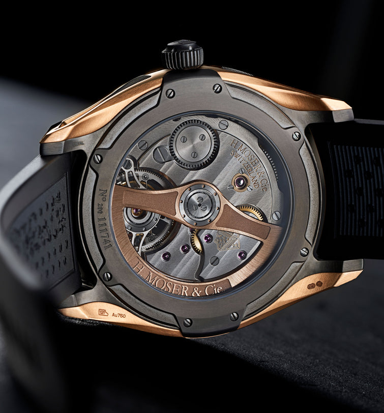Tourbillon watch with rose gold and gunmetal case, visible mechanical movement and exposed balance wheel, black rubber strap.