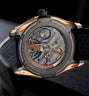 Tourbillon watch with rose gold and gunmetal case, visible mechanical movement and exposed balance wheel, black rubber strap.