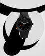 Black watch with minimalist dial, silver hands, orange accent detail on side, and black textile strap.
