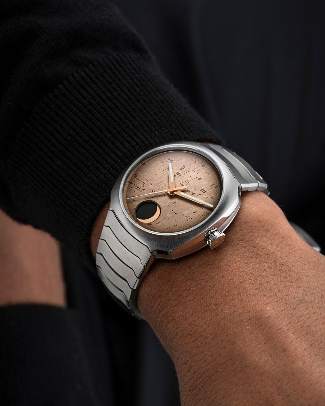 A close-up of a person with medium skin tone wearing an H. Moser & Cie Streamliner Perpetual Moon Concept Meteorite, featuring a sleek silver case, textured brown dial, rose gold hands, and a black accent, paired with a black long-sleeve shirt.