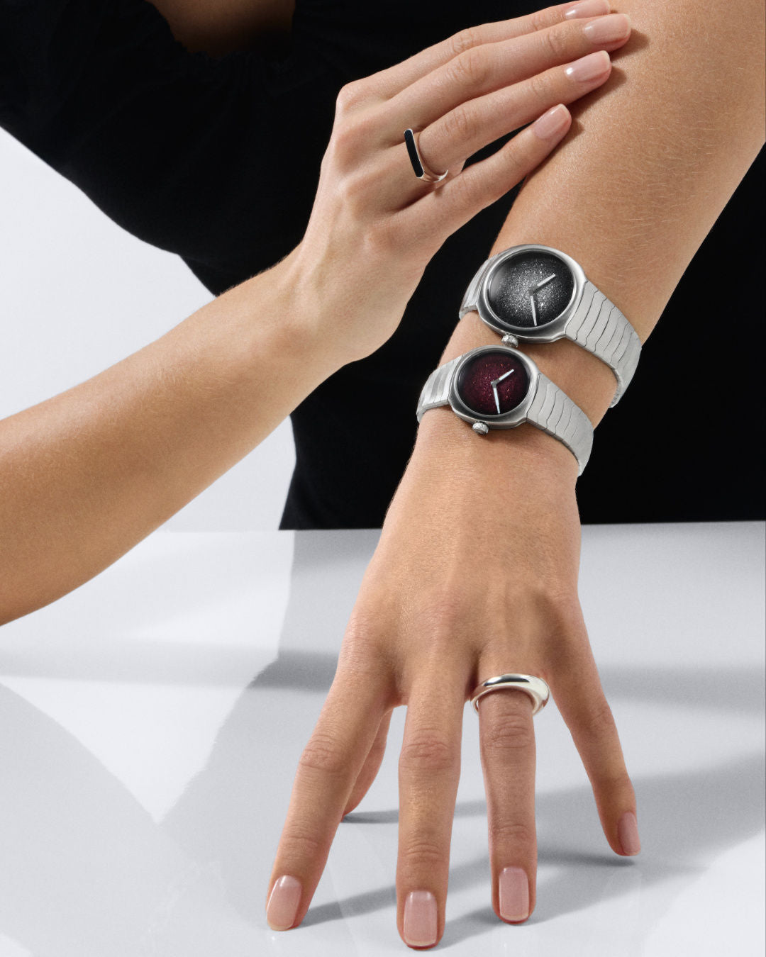 Two wristwatches with white metal bracelets worn on one wrist, featuring black and burgundy dial faces with minimalist design