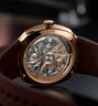 Rose gold watch with visible tourbillon mechanism, skeleton dial showing gears and components, brown leather strap, 12 ATM wa