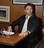 Man wearing black leather jacket and white shirt seated at wooden table, wristwatch visible on left wrist.