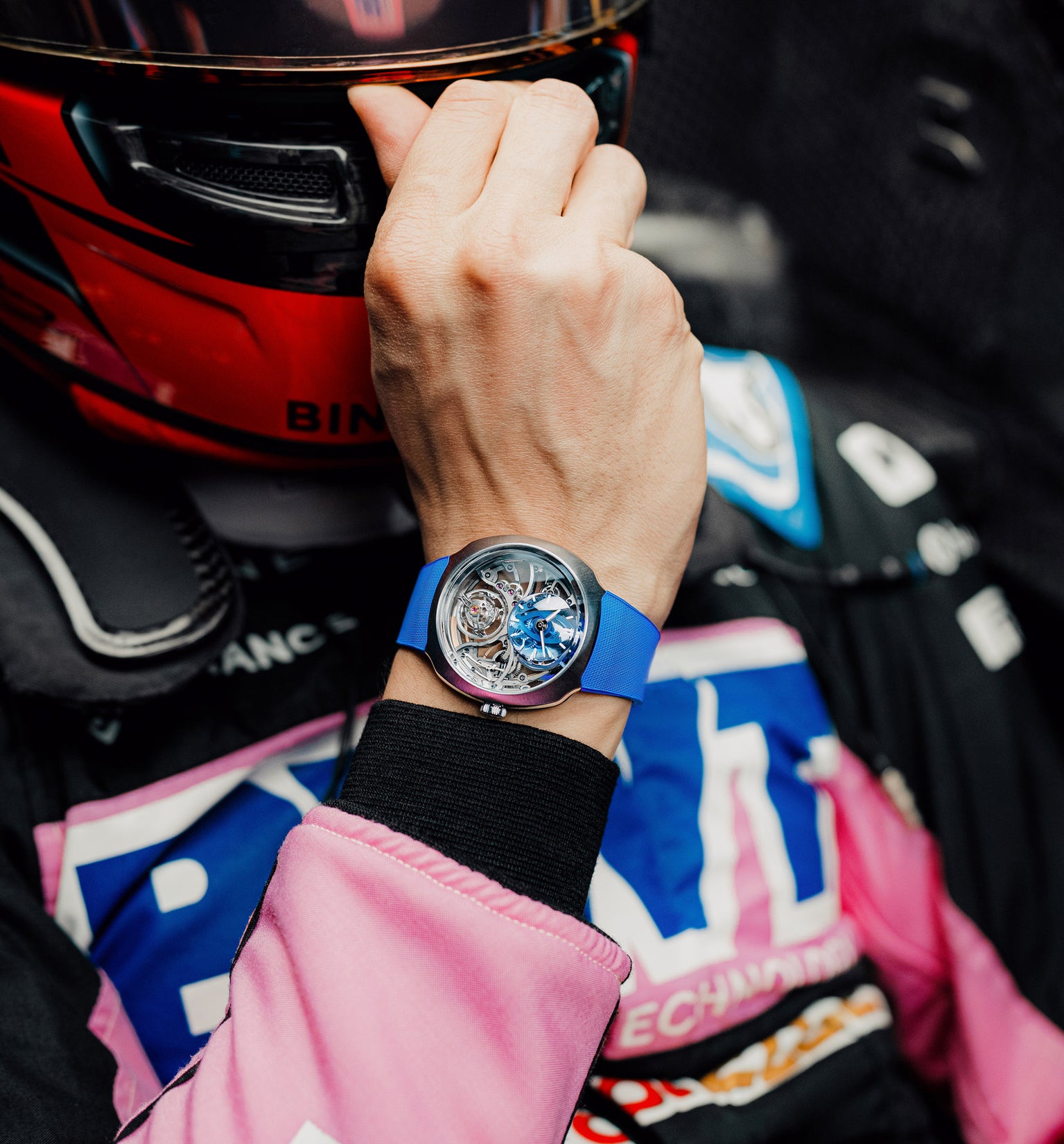 Wrist watch with blue rubber strap and silver case worn on wrist holding red and black helmet.