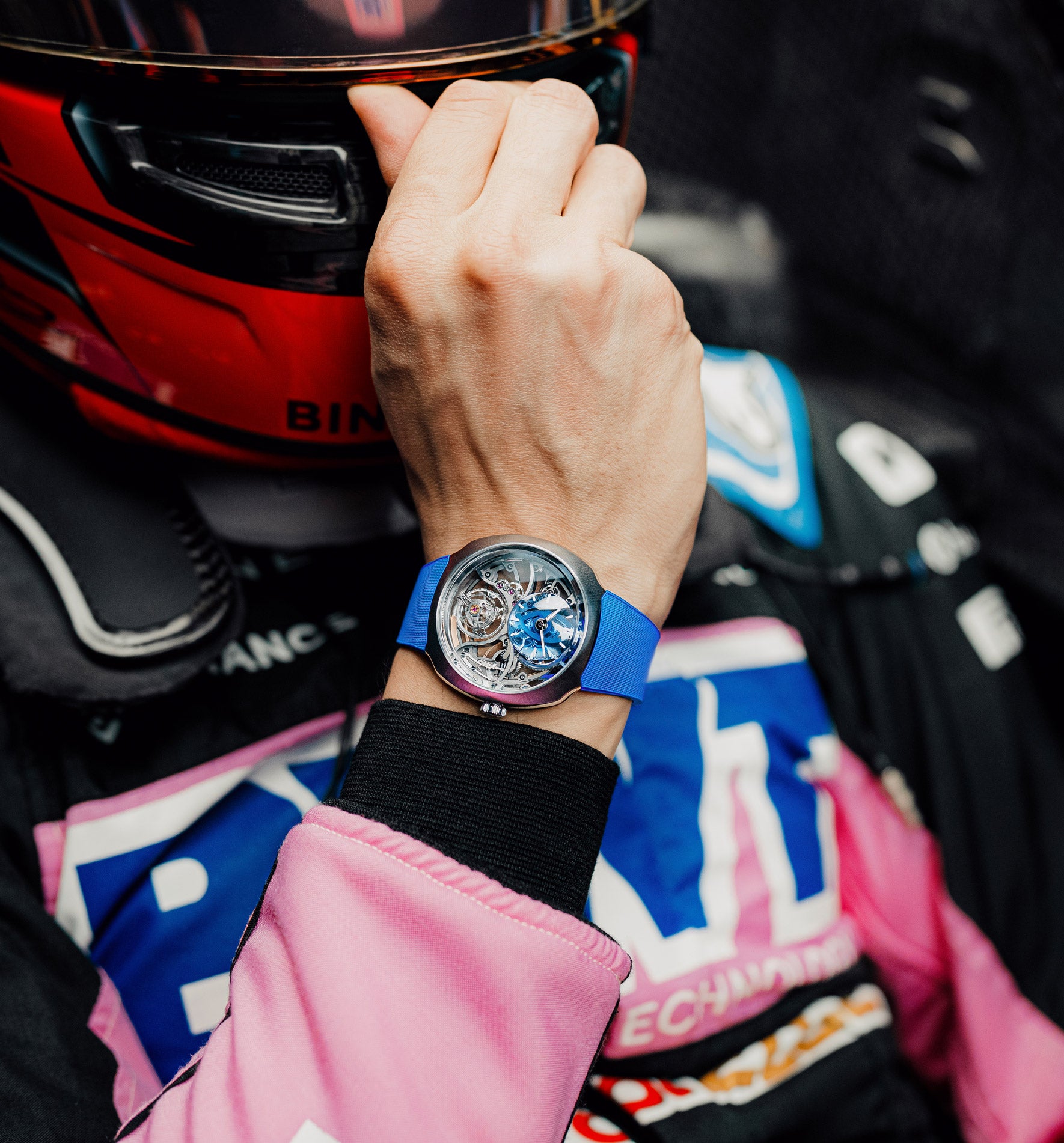 Wrist watch with blue rubber strap and silver case worn on wrist holding red and black helmet.