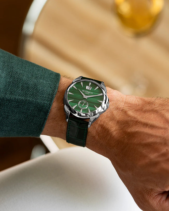 Men's watch with green dial, silver case, black leather strap worn on wrist with green sleeve visible.