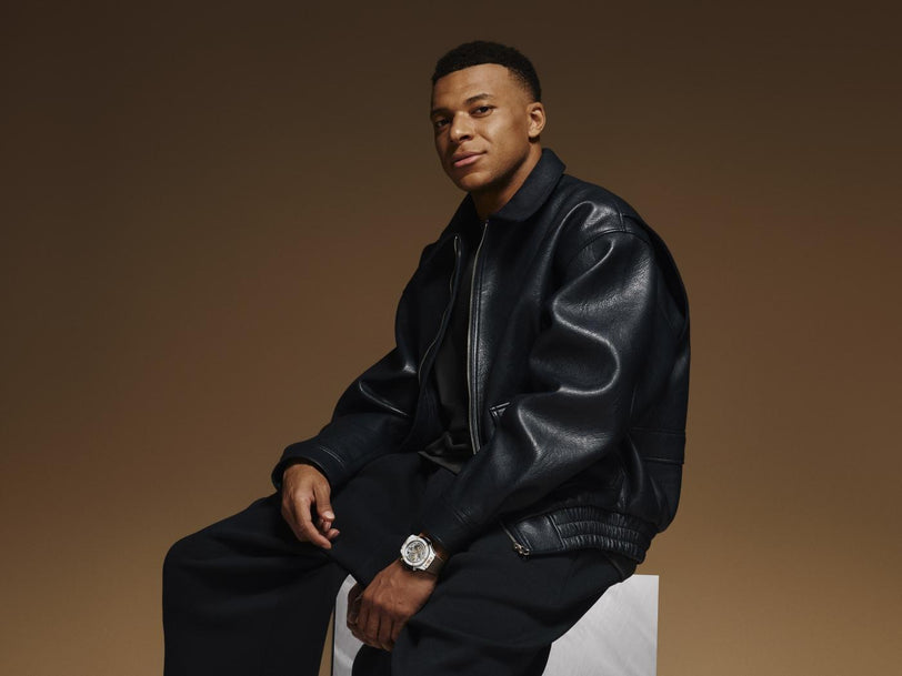 Man wearing black leather jacket and black pants with white ceramic watch on wrist against tan background.