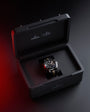 Black and red Omega presentation box with open lid displaying chronograph watch with black dial, red bezel, and leather strap