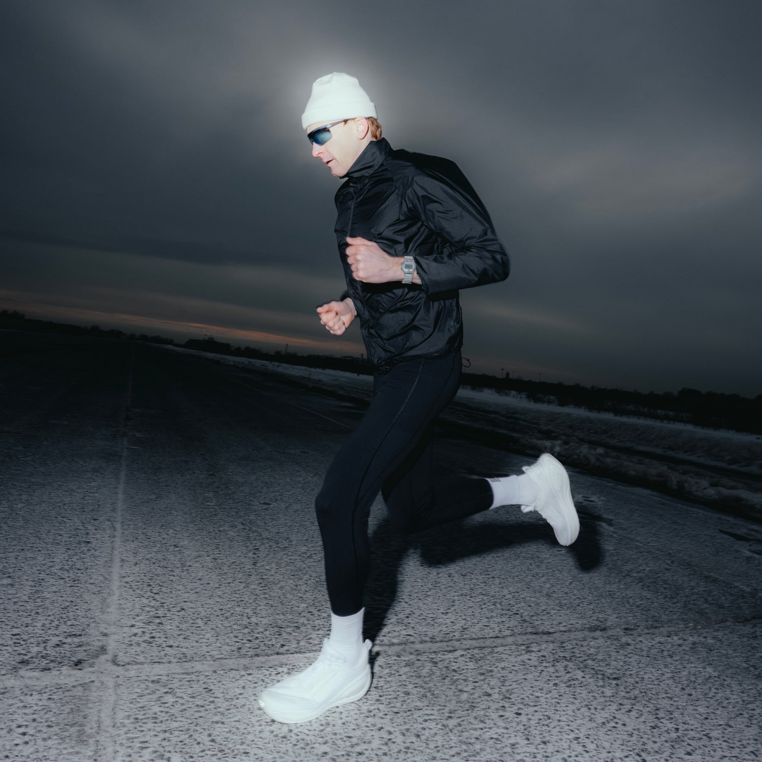 Person jogging at dusk wearing Timex Ironman watch on left wrist, black jacket, white beanie, and sunglasses.