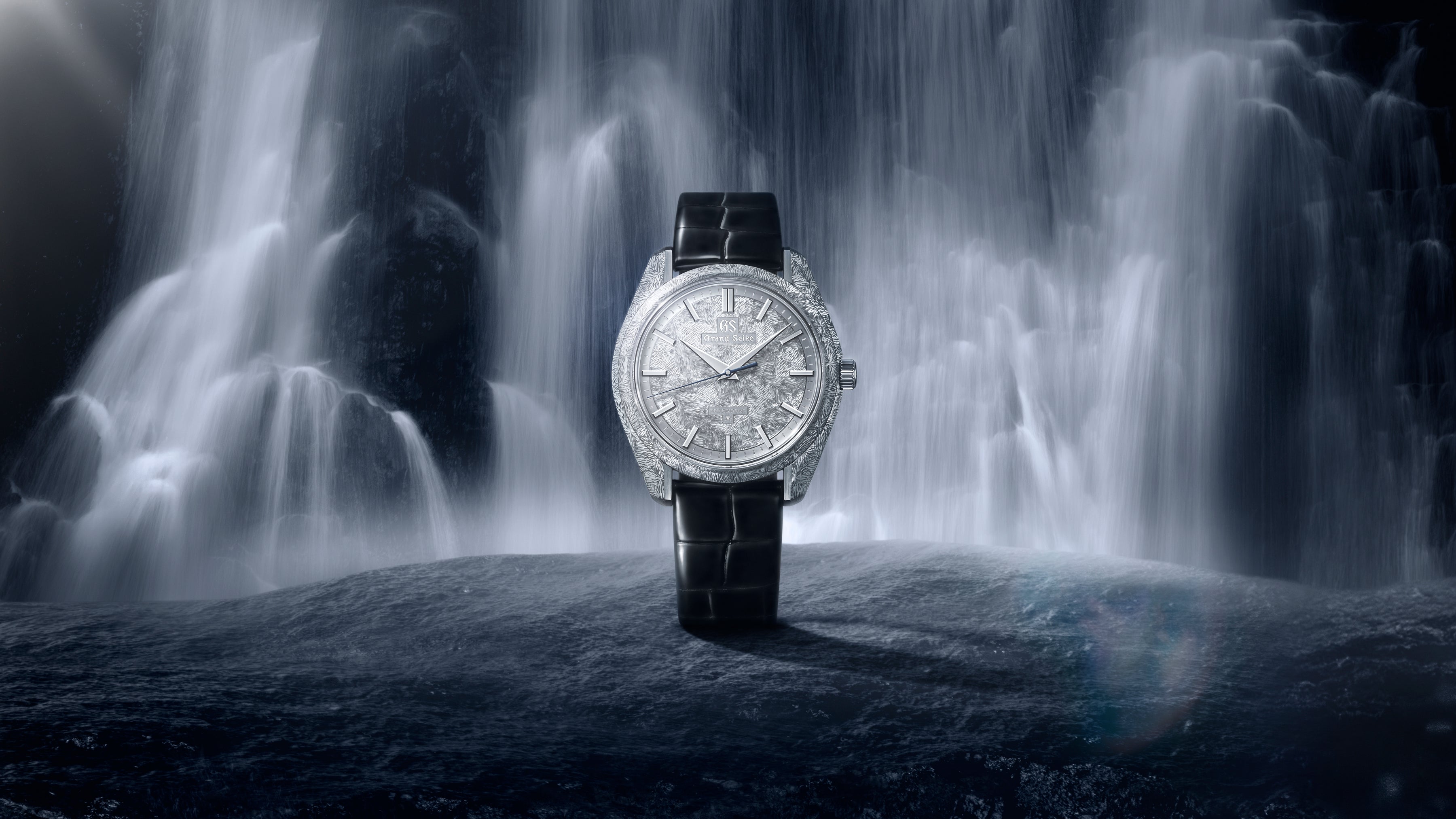 Silver-dial watch with black leather strap displayed against a waterfall backdrop.