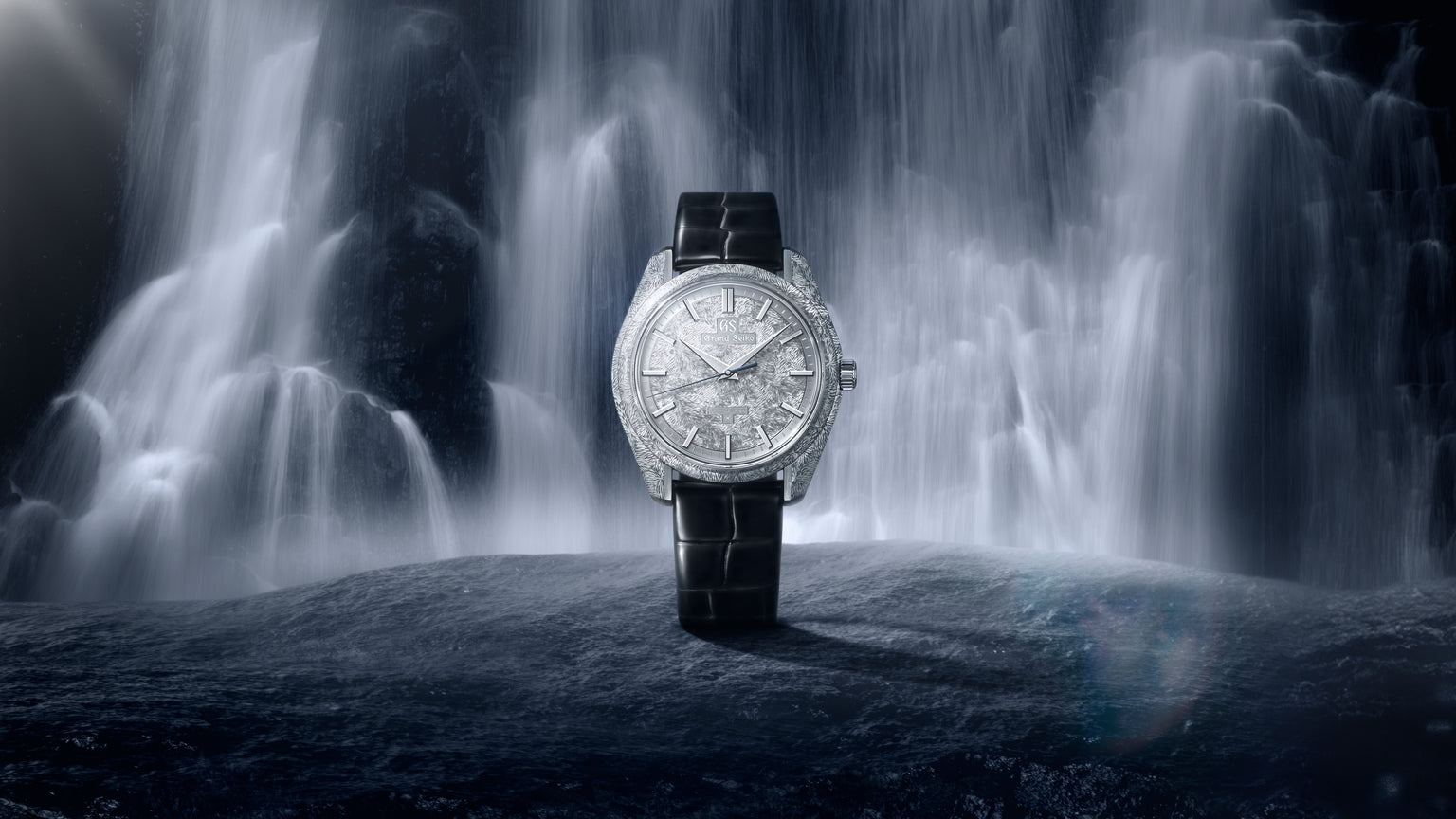 Silver-dial watch with black leather strap displayed against a waterfall backdrop.