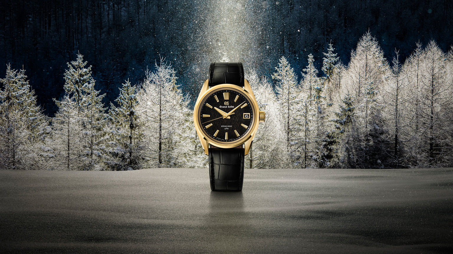 Gold-tone watch with black dial, black leather strap, and date window against a frost-covered forest backdrop.