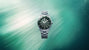 Steel diver watch with black dial, luminous hour markers, rotating bezel, and matching steel bracelet against teal water back