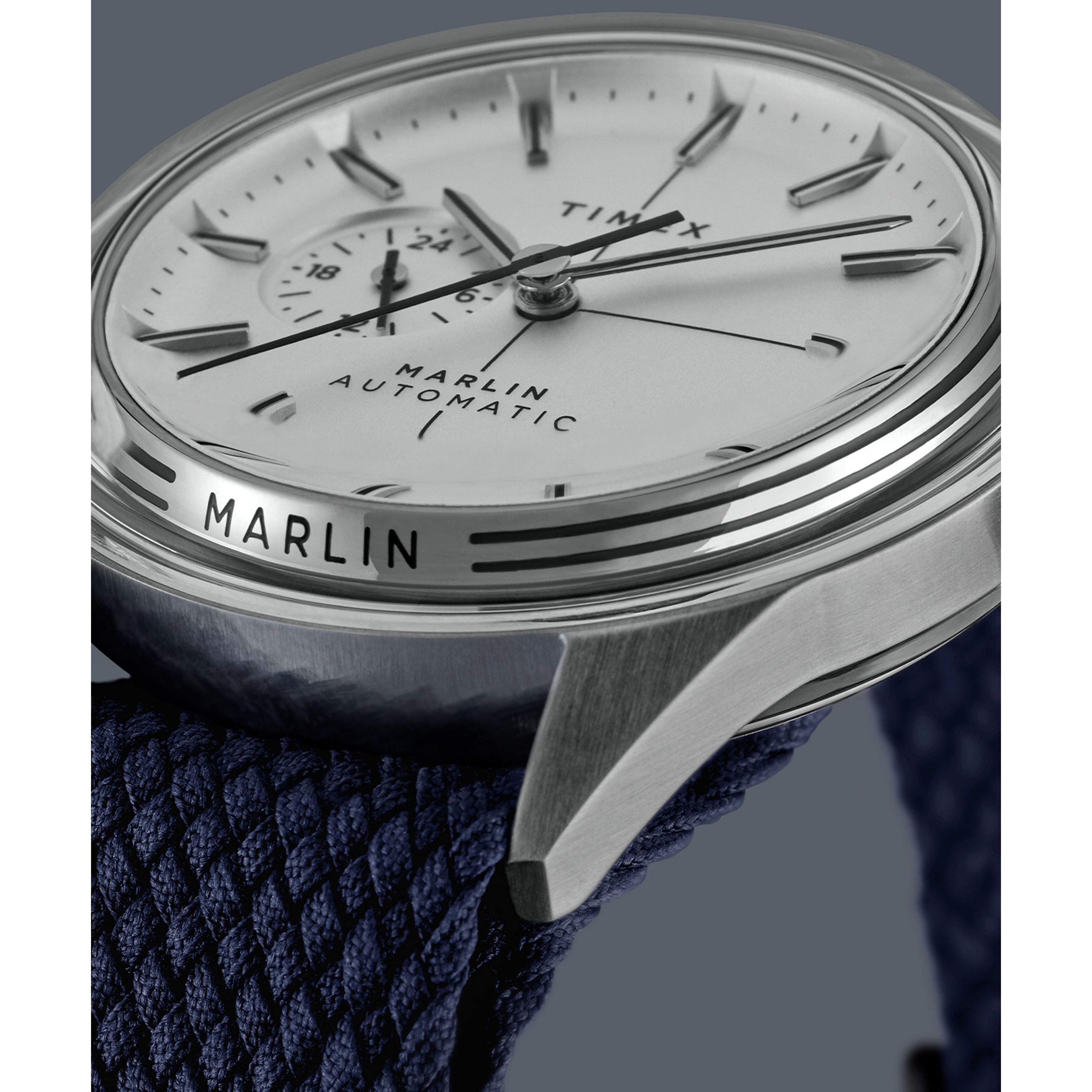 Close-up of a Timex Marlin® Jet Automatic wristwatch, showcasing a domed Hesalite crystal over a white dial with black hour markers and a blue textured strap. The words MARLIN® JET AUTOMATIC and TIMEX are visible on its retro-futuristic design.
