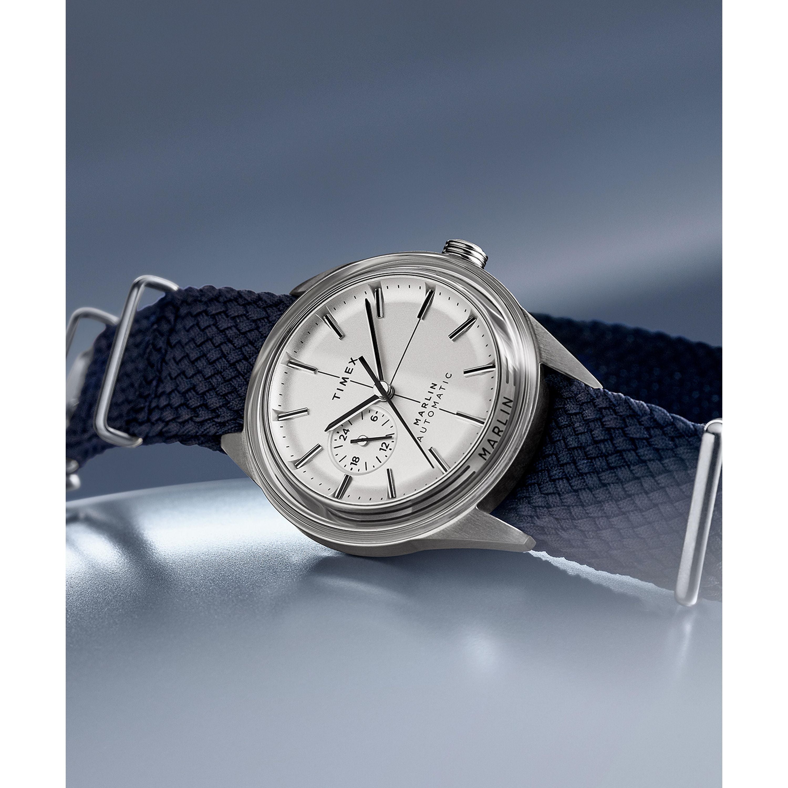 The Timex Marlin® Jet Automatic features a retro-futuristic silver case, white dial with black hour markers, small subdial, and textured navy blue strap under a domed Hesalite crystal, set against a soft blue gradient background.