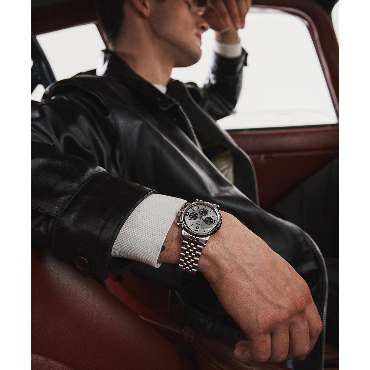 A person in a black leather jacket and sunglasses sits in a car, leaning back with one arm on the door, showcasing a Timex Marlin® Chronograph Tachymeter watch with sleek quartz movement on their wrist.