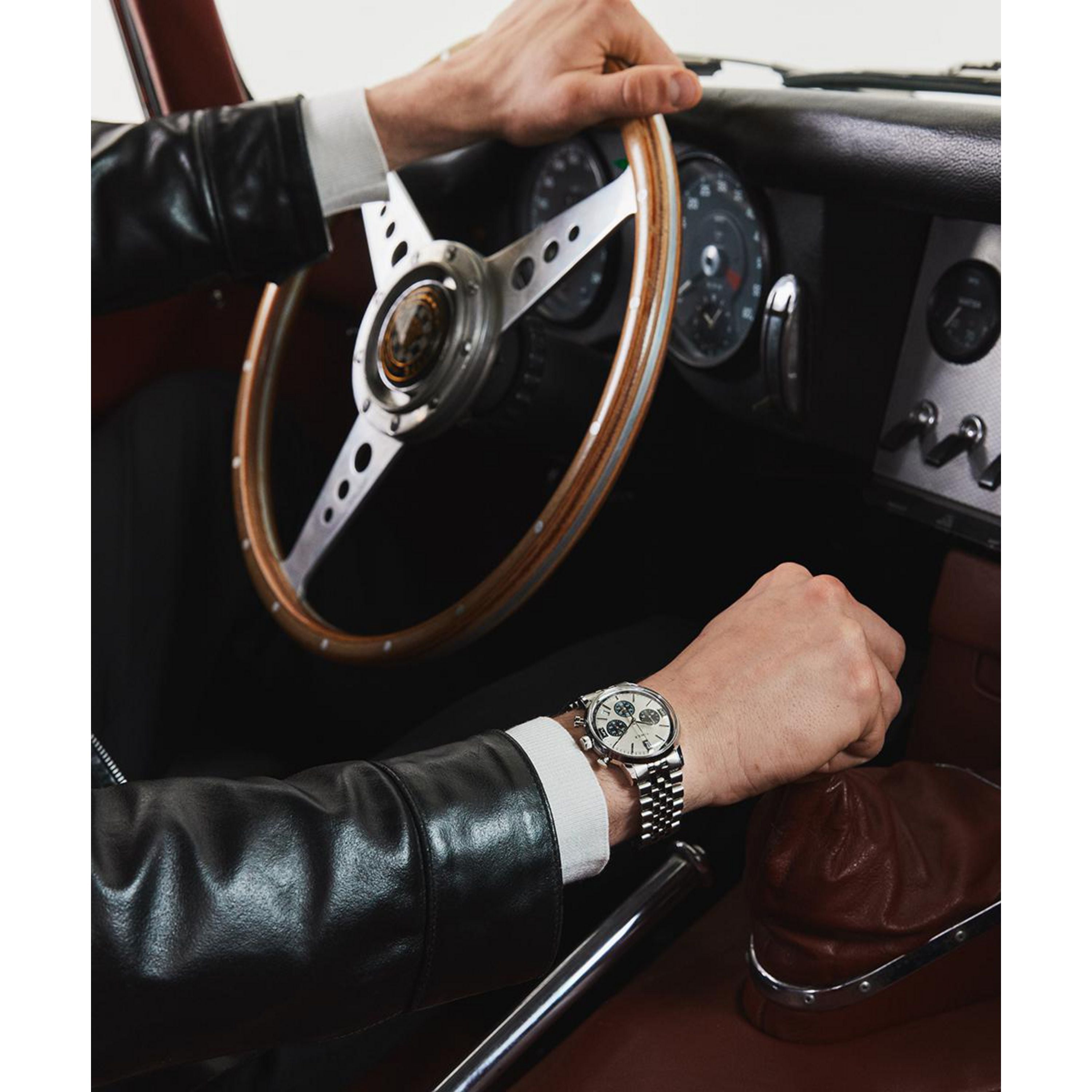 Wearing a black leather jacket, a driver grips a vintage car's wooden steering wheel with one hand and the gear shift with the other, sporting a Timex Marlin® Chronograph Tachymeter with a sleek silver bezel.
