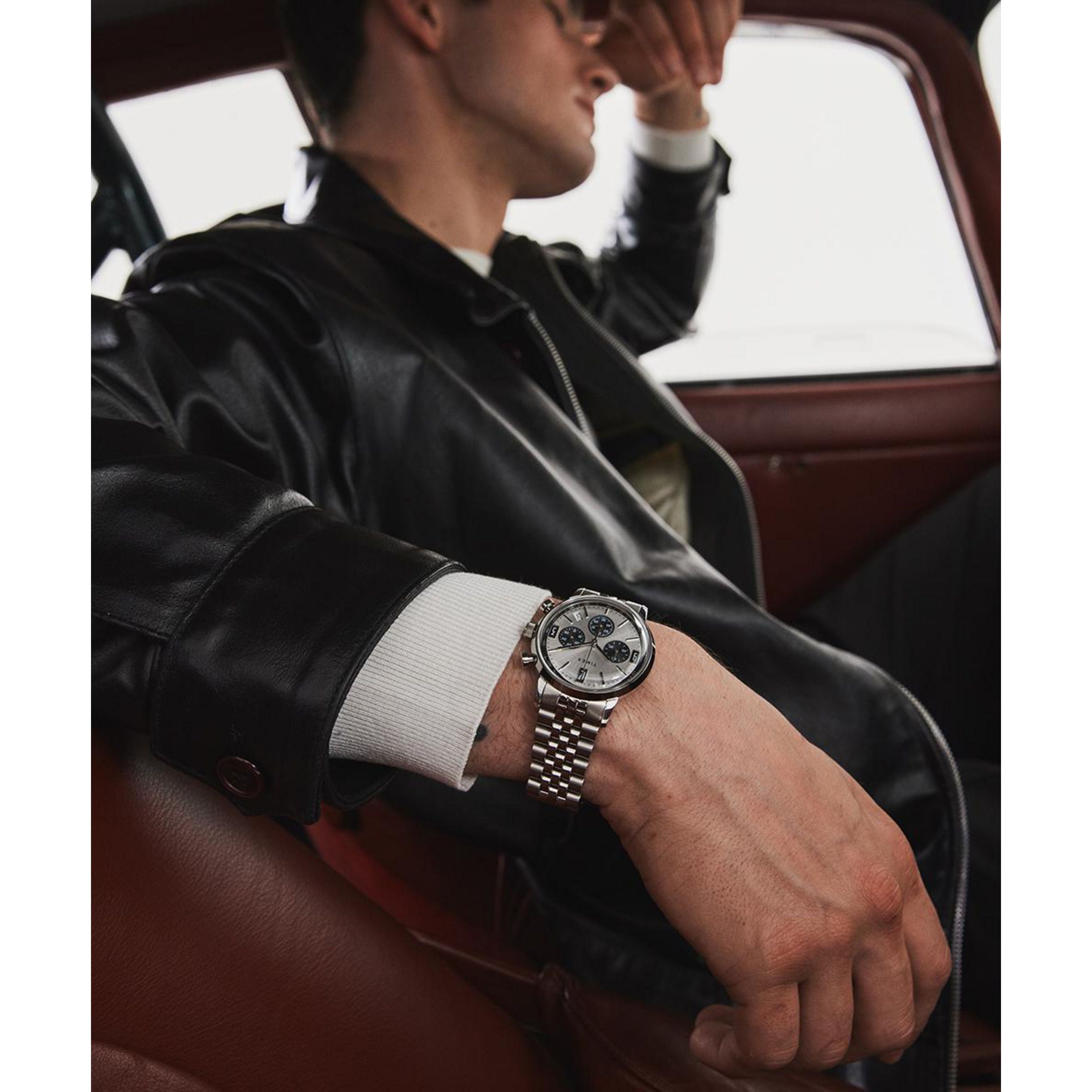 A man in a black leather jacket sits in a car, his left arm on the seat, wearing a Timex Marlin® Chronograph Tachymeter wristwatch with a metal band and white dial. He looks out the window, shielding his eyes with his hand.