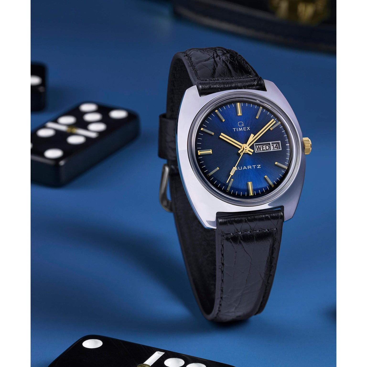 A Timex G SS QZ SW 1978 DAY DTE 37.5 BLU DL 25 watch with a black leather strap and blue sunray dial showing day and date, displayed on a blue surface with domino pieces around it.