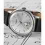A Timex x Peanuts Marlin® Automatic Saxophonist watch by Timex features a silver dial, black leather strap, and Snoopy as a saxophonist on the face, displayed atop a surface decorated with Snoopy comic strip illustrations.