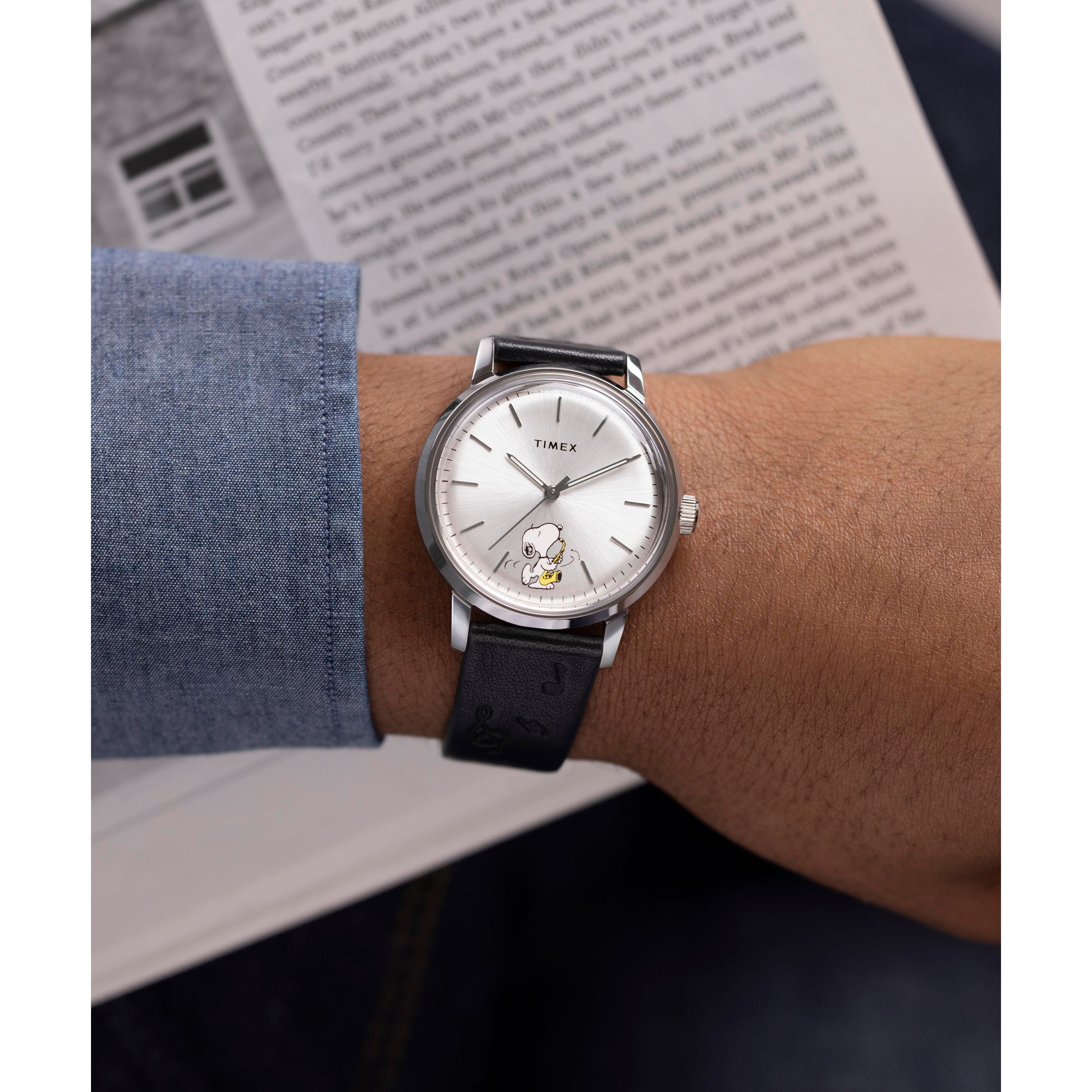 A person in a blue denim shirt holds an open book while wearing the Timex x Peanuts Marlin® Automatic Saxophonist watch, featuring a silver case, black leather strap, and saxophone-themed dial by Timex.