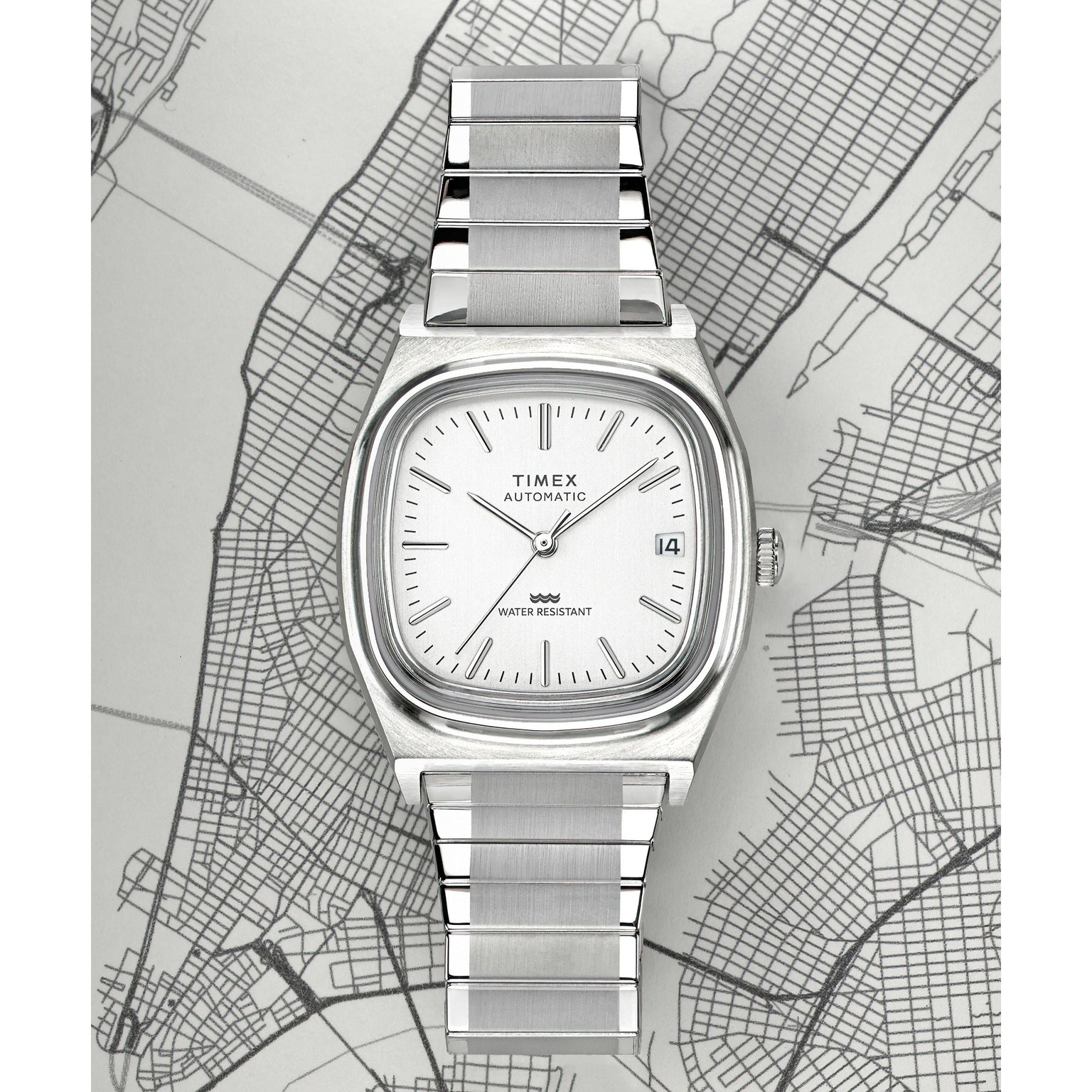 The Timex G SS AU BW E LINE REISSUE 34 WHITE DL 25 wristwatch, with a silver metal band and white dial, is shown on a grayscale city map. It features simple markers, a date at 3 o’clock, and Japanese automatic movement.