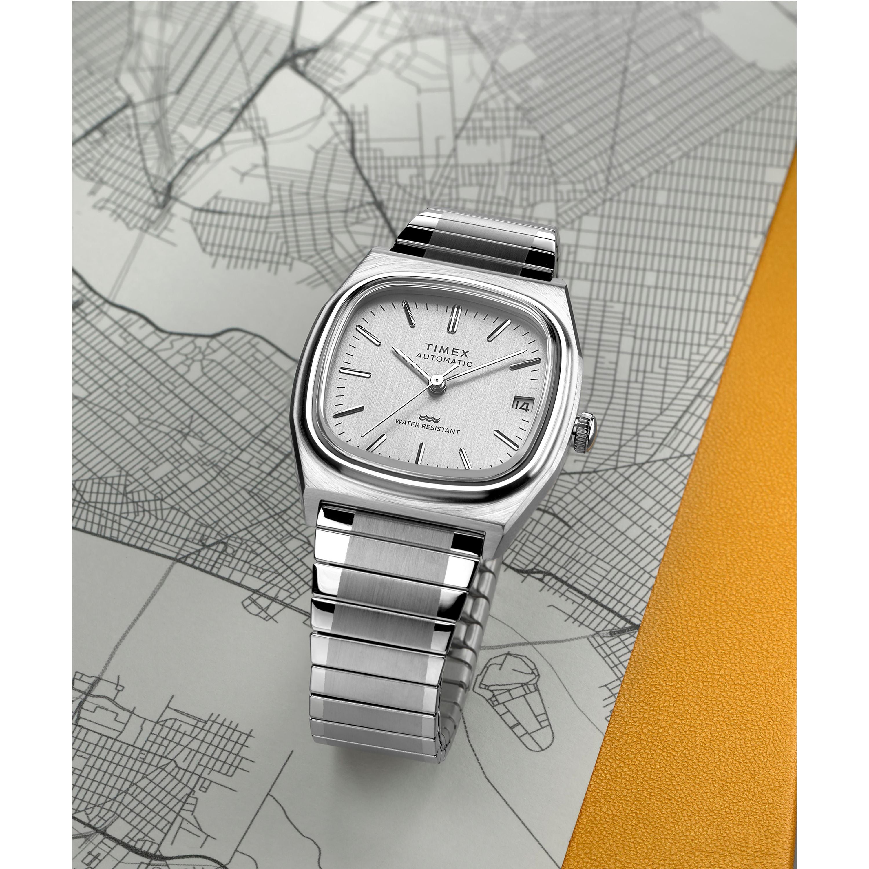 The Timex G SS AU BW E LINE REISSUE 34 WHITE DL 25 wristwatch rests on a grayscale city map with a yellow section. Set to 10:09, it features Japanese automatic movement, date display, and a silver square TV dial with metal band.
