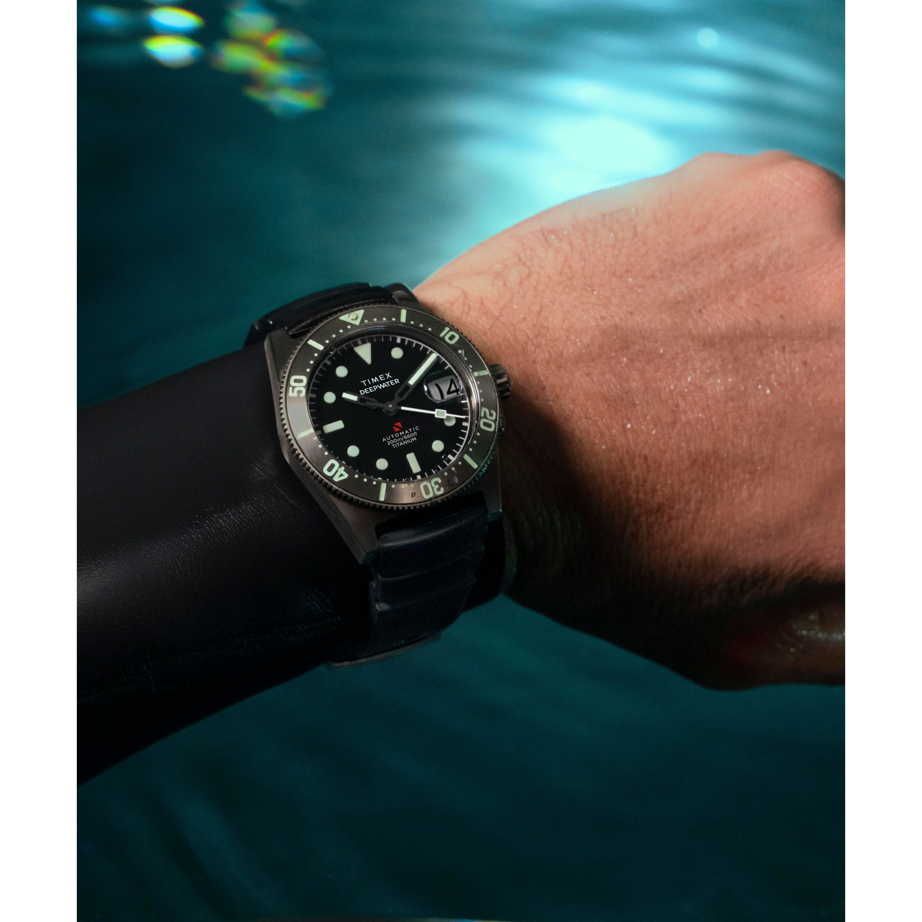 A diver showcases the Timex G TI AU SW DEEP WTR REEF 41 BLAK DL 25—a titanium automatic dive watch with black bezel and strap—above shimmering blue water.