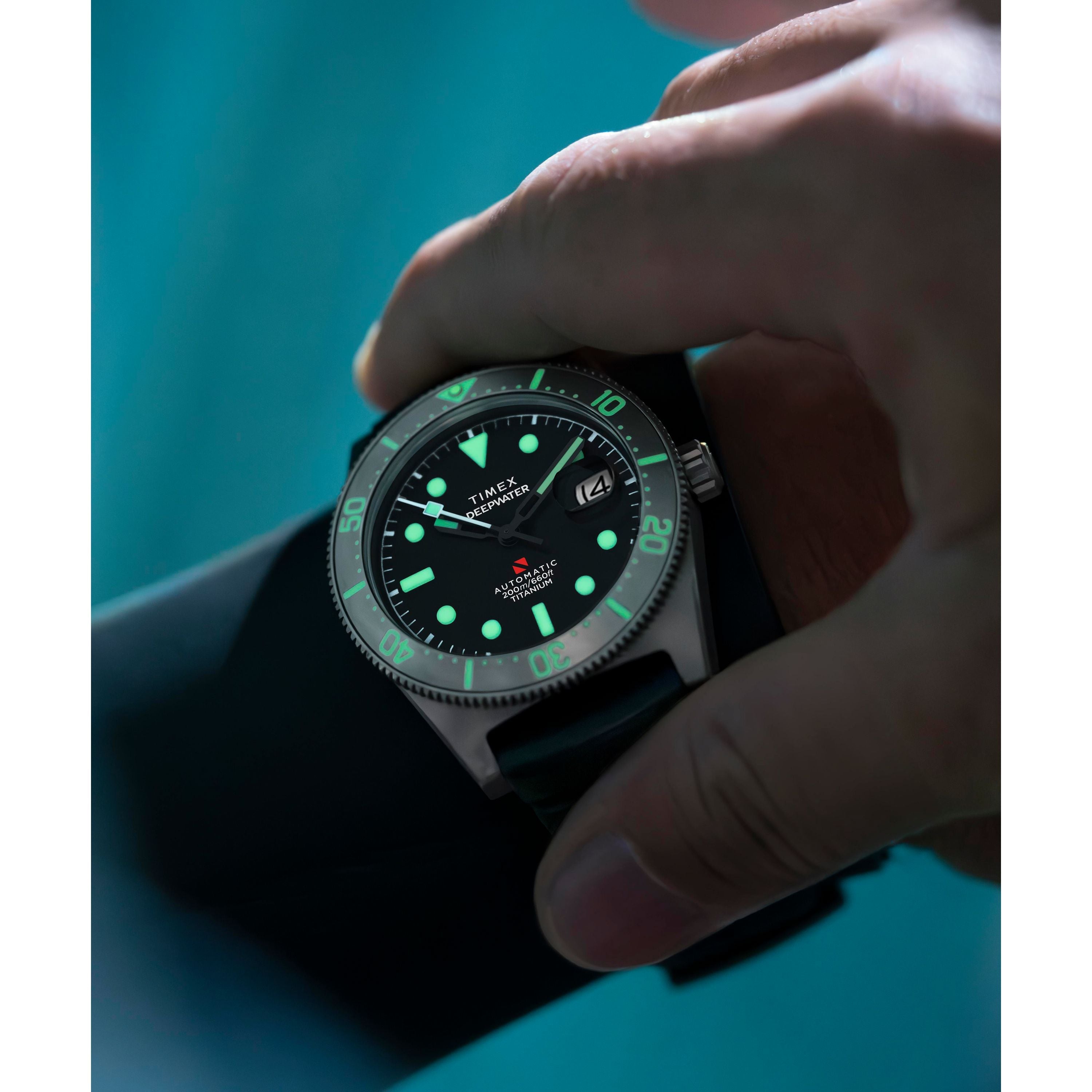 A close-up of a hand wearing the Timex G TI AU SW DEEP WTR REEF 41 BLAK DL 25 titanium automatic dive watch with a sapphire crystal, luminous dial, clear time and date display, set against a blurred blue background.