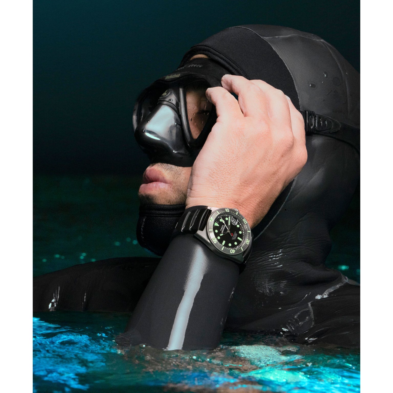 A person in a black wetsuit and diving mask, partially submerged, adjusts their mask. On their wrist, the Timex G TI AU SW DEEP WTR REEF 41 BLAK DL 25 titanium automatic dive watch with green dial and 200m water resistance stands out.