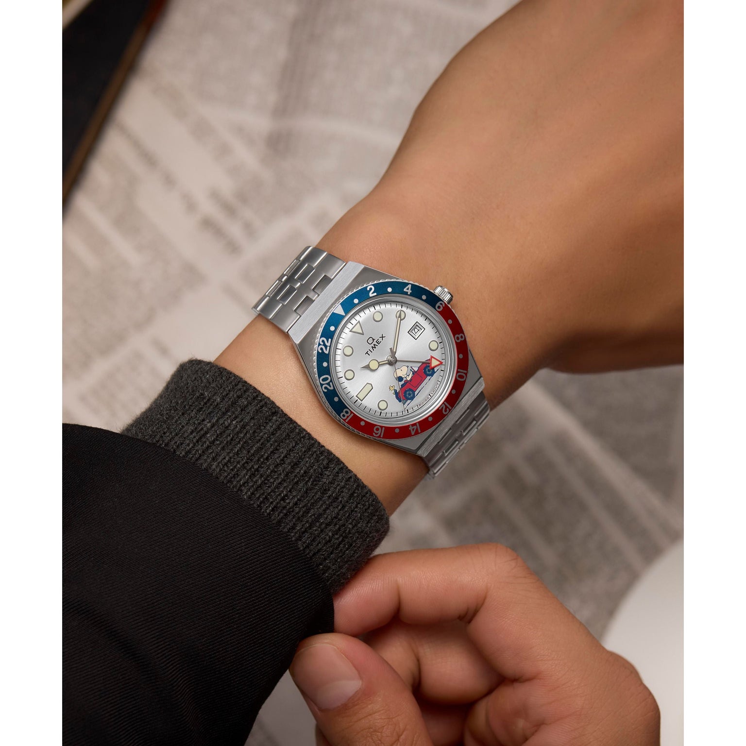 A person wears the Timex G SS QZ PEANUTS GMT 38 SLVR DL SS BLT 25, a stainless-steel watch with a red and blue bezel and white dial, on their left wrist over a dark sleeve; a blurred newspaper is in the background.