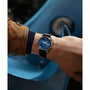 A person in a brown coat and navy sweater showcases the Timex Marlin® Automatic wristwatch, featuring a blue face and eco-friendly leather strap; a curved blue car with visible headlight is seen in the background.