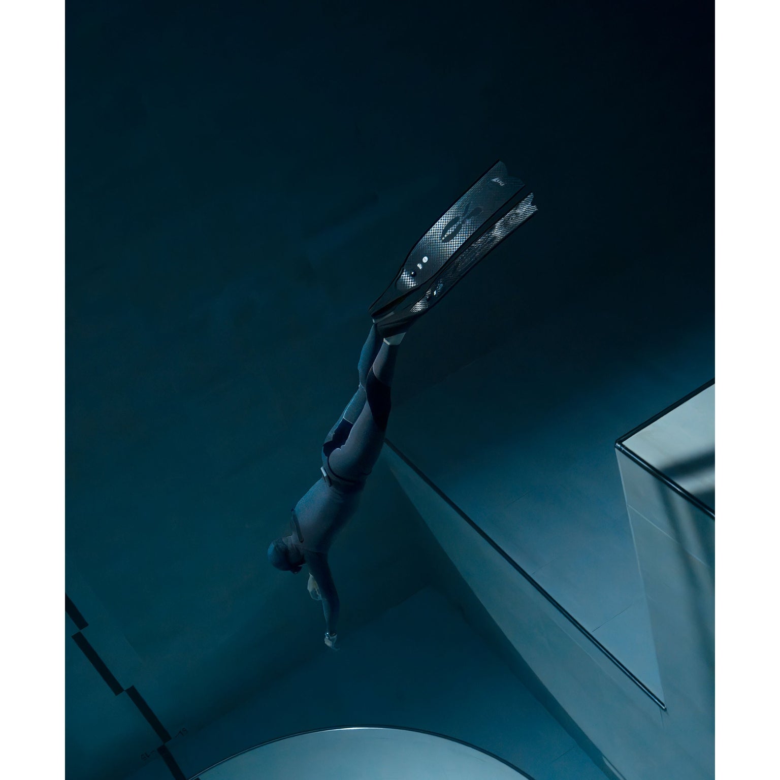 A person in a wetsuit and fins swims underwater in a deep blue pool, wearing the Timex Deepwater Reef 200 dive watch with 200m water resistance. The calm, minimalistic scene features soft lighting and geometric pool edges.