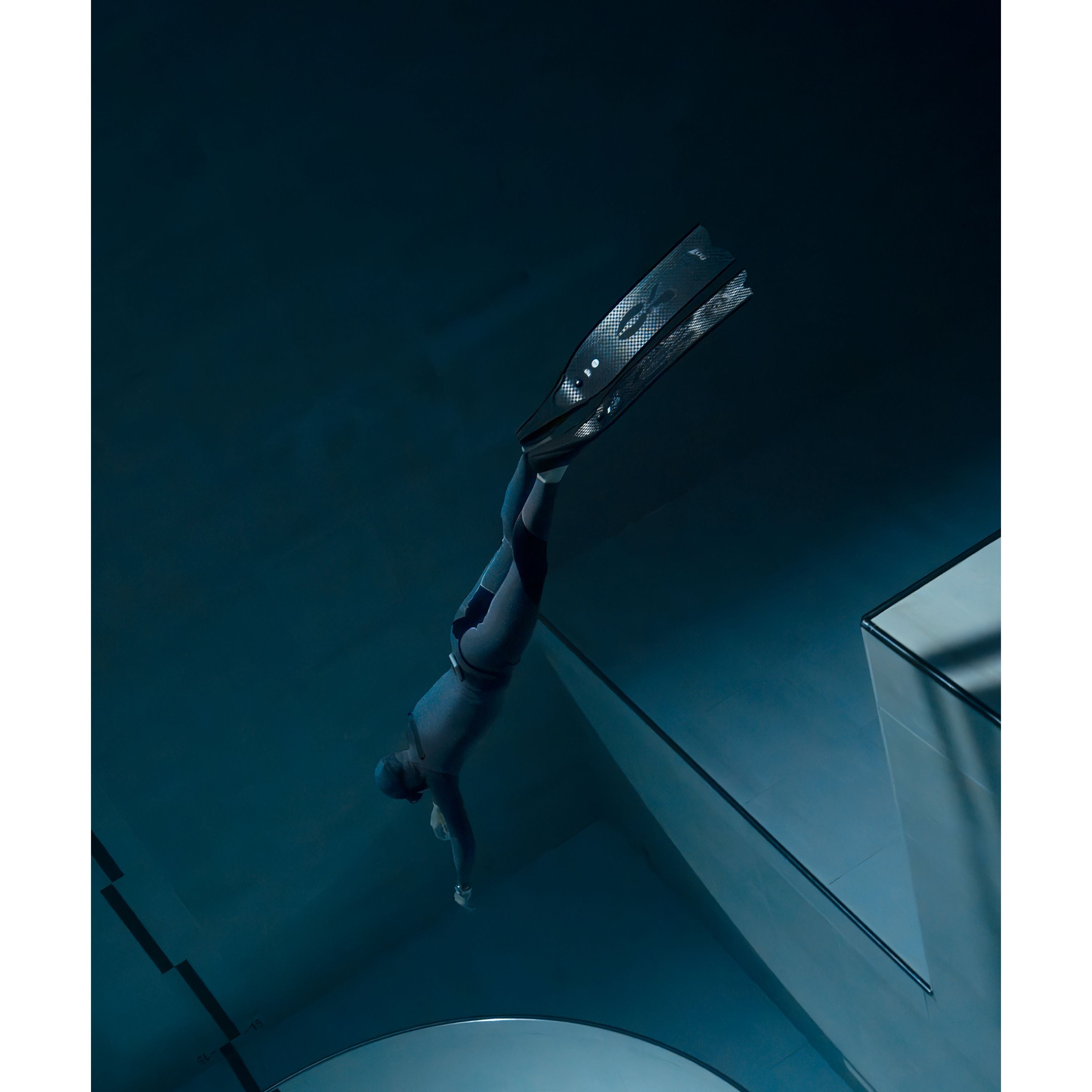 A person in a wetsuit and fins swims underwater in a deep blue pool, wearing the Timex Deepwater Reef 200 dive watch with 200m water resistance. The calm, minimalistic scene features soft lighting and geometric pool edges.