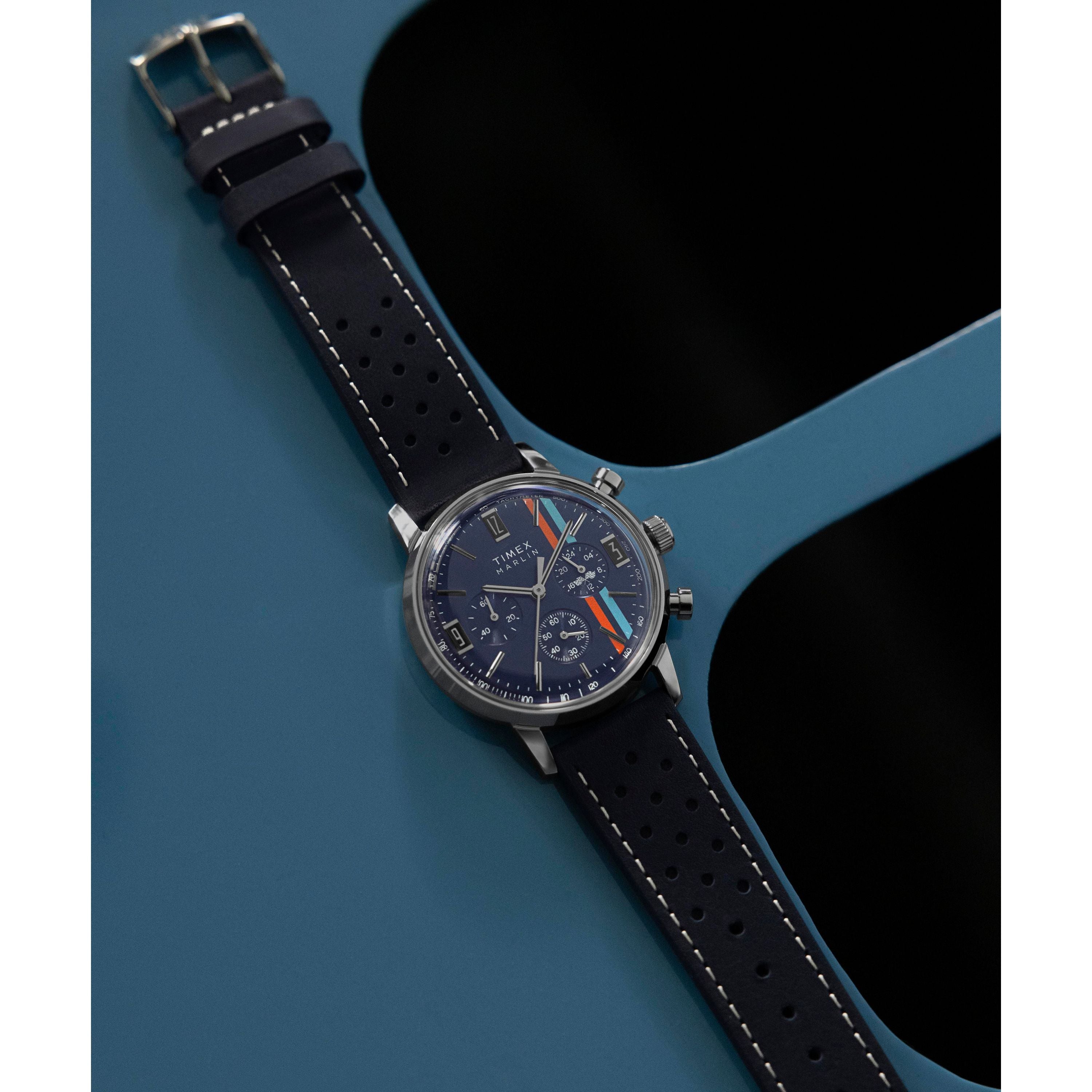 The Timex Marlin® Chronograph Tachymeter features a dark blue face with racing stripes and multiple dials, resting diagonally on blue. Its black leather strap with white stitching adds timeless style to this bold Marlin chronograph.
