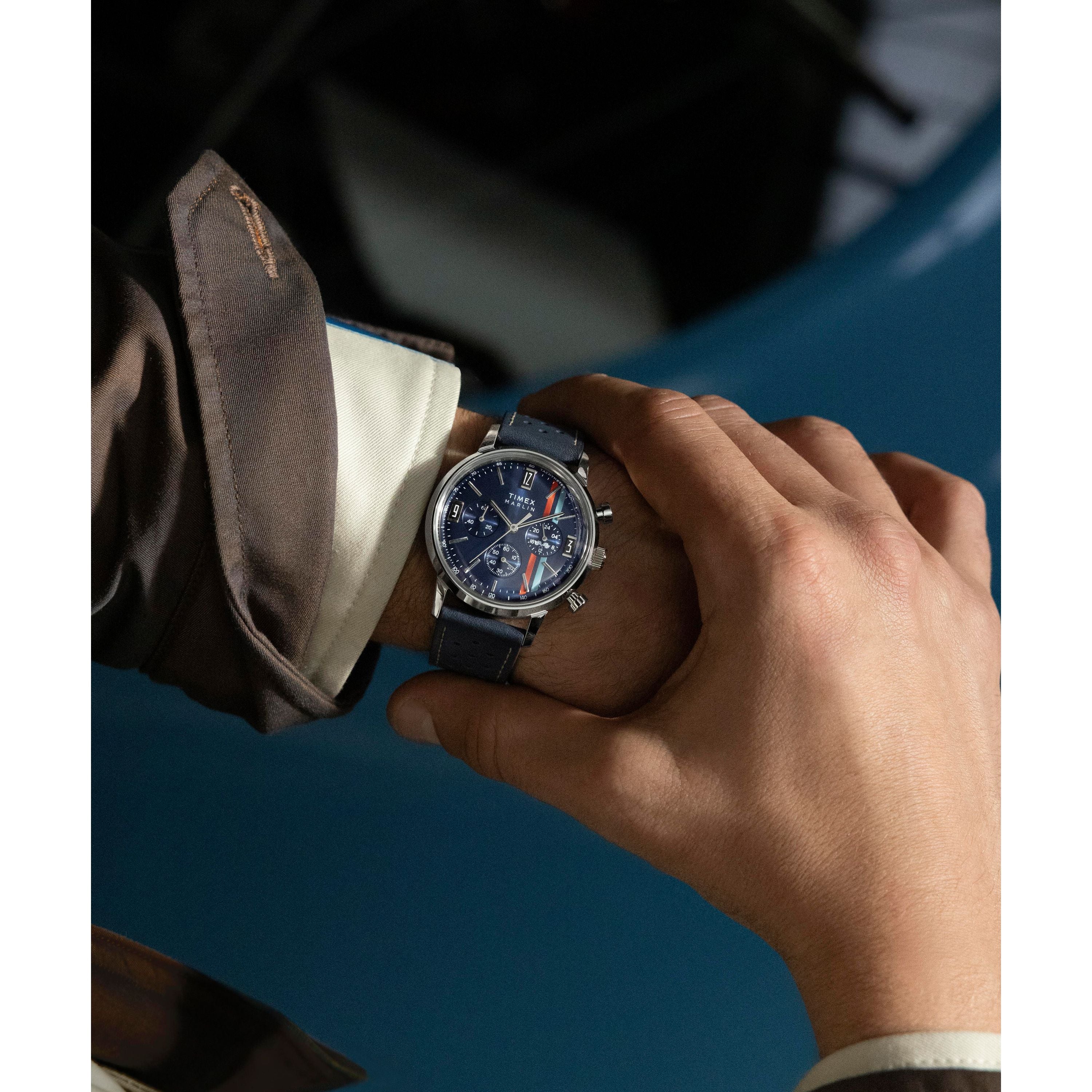 A person wearing a Timex Marlin® Chronograph Tachymeter with a leather strap on their left wrist adjusts it with their right hand. They're dressed in a brown jacket over a white shirt, with a blue surface in the background.
