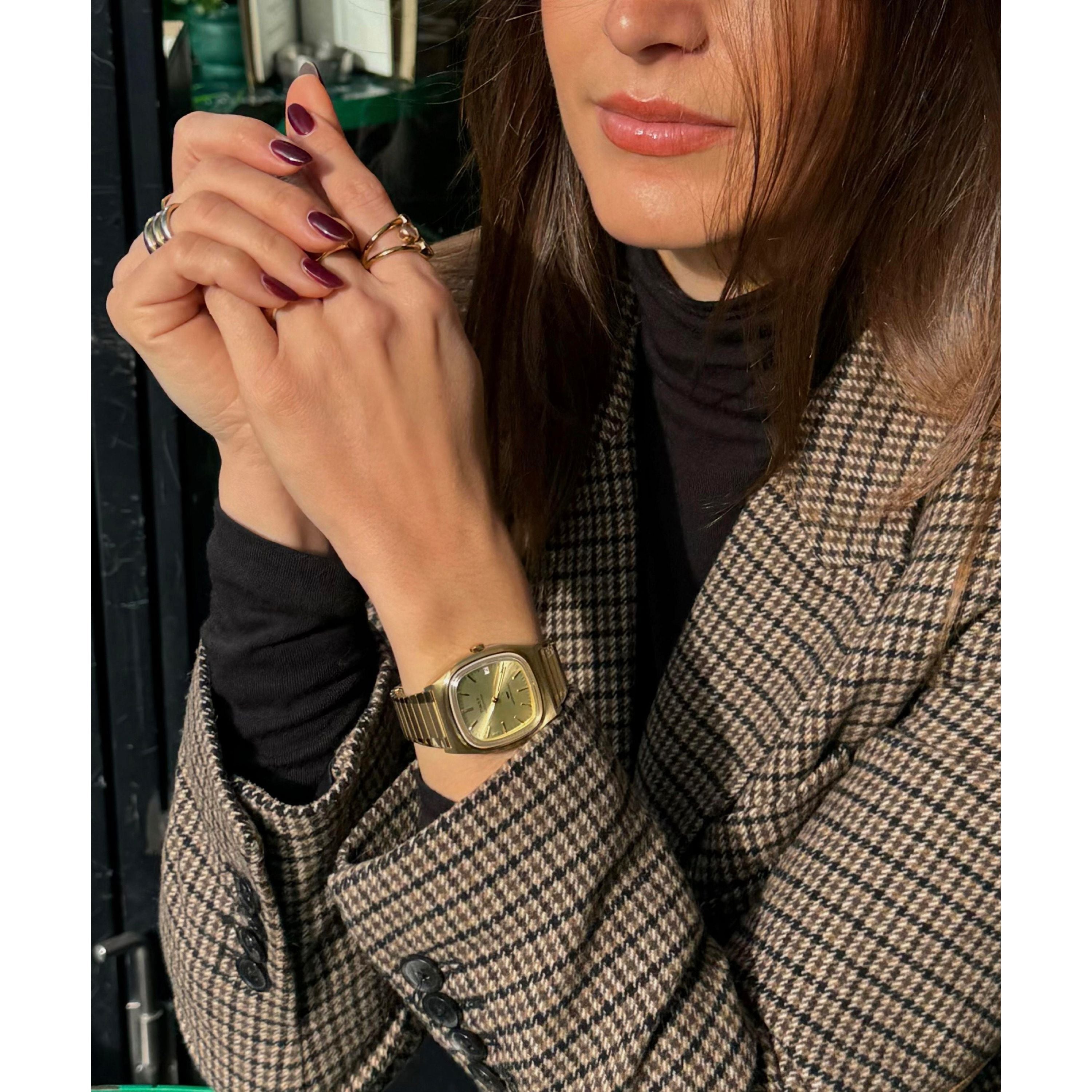 A woman in a brown plaid blazer and black turtleneck clasps her hands, displaying burgundy nails, gold rings, and a Timex Automatic 1983 E Line Reissue watch. Sunlight highlights her face and accessories.