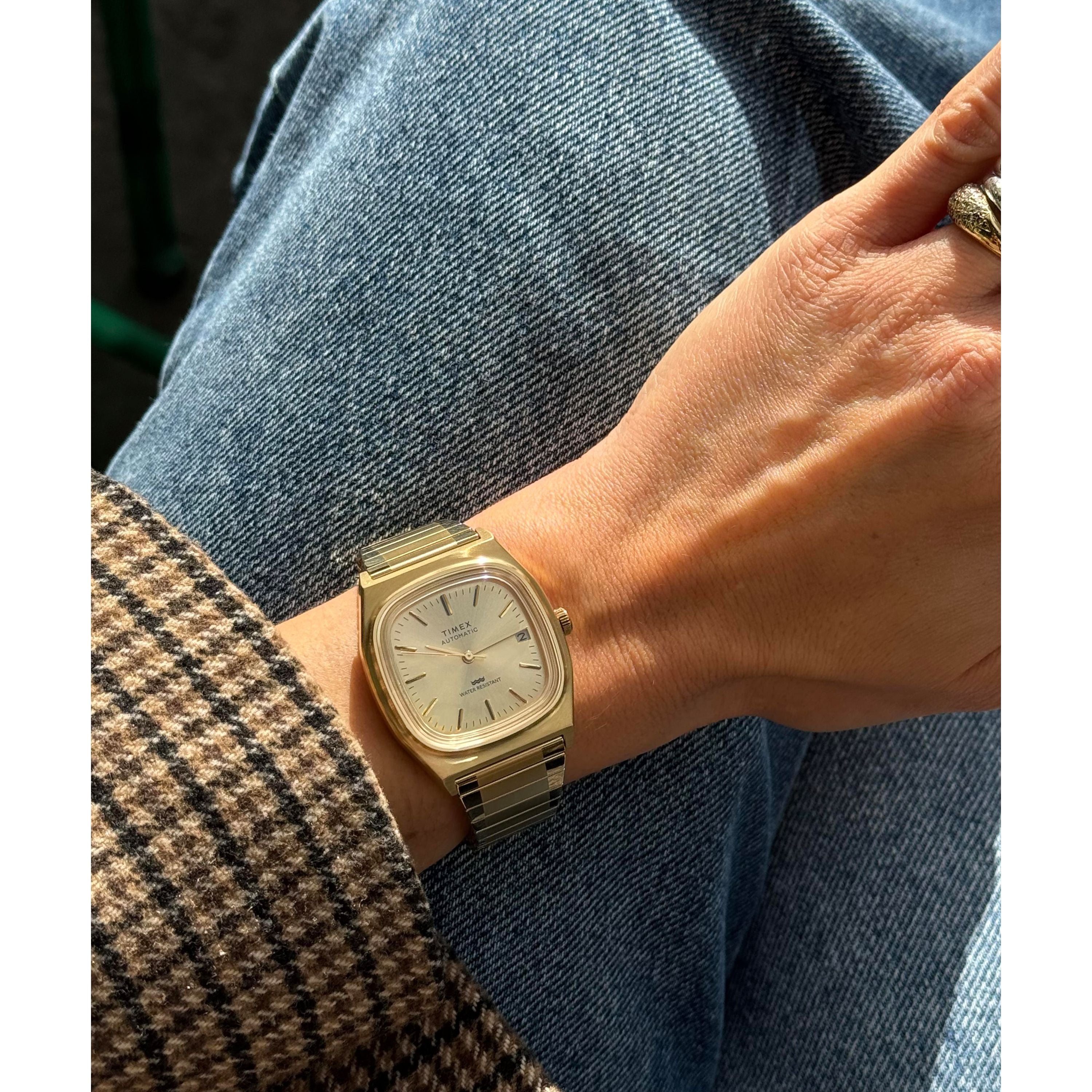 A person wearing a Timex Automatic 1983 E Line Reissue gold-tone dress watch sits with crossed legs, blue jeans, and a brown houndstooth coat. Sunlight highlights the watch’s Japanese automatic movement and their ring-adorned hand.