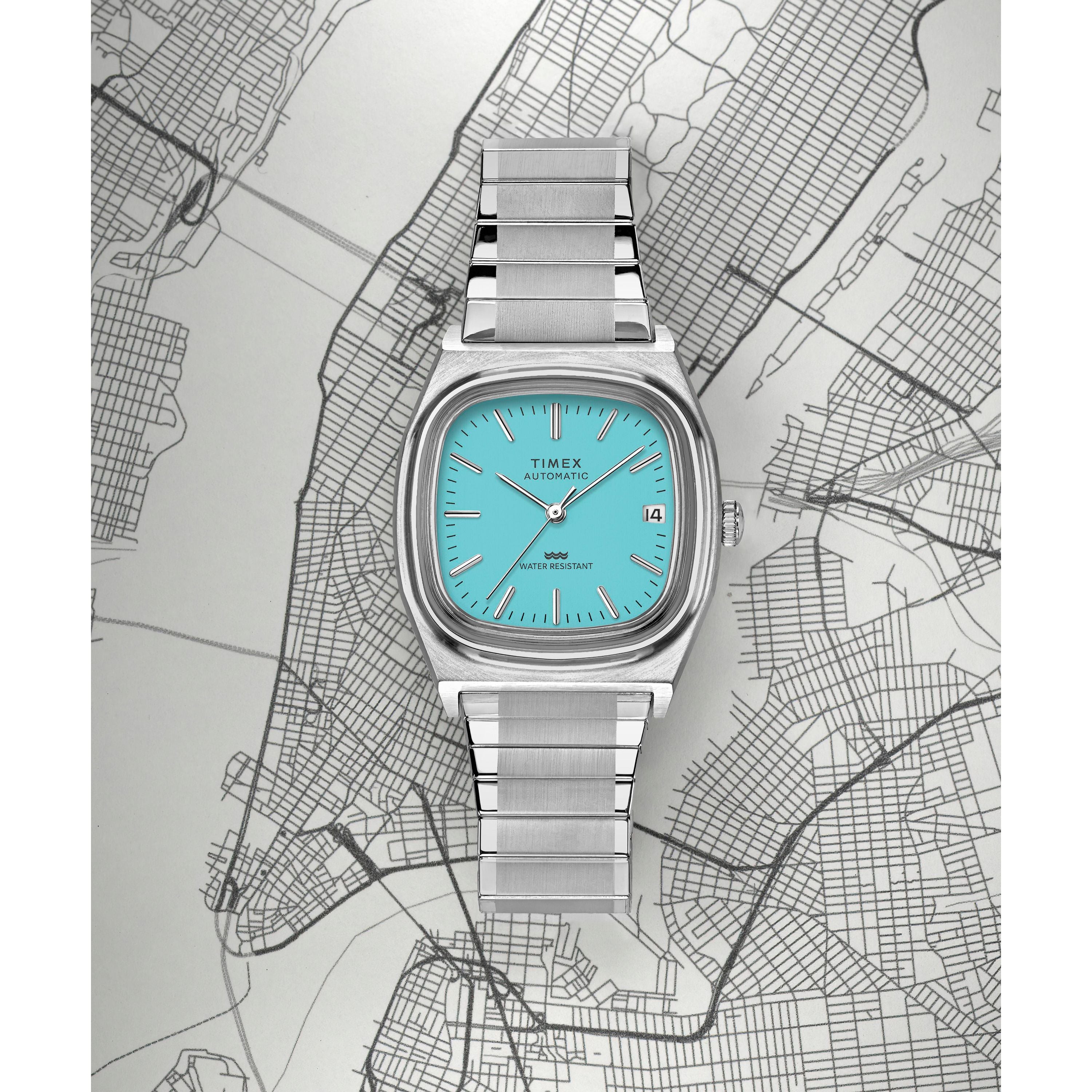 A Timex Automatic 1983 E Line dress watch with a turquoise face and metal band is displayed on a black-and-white New York City map, showing the date and time with the cityscape visible beneath it.