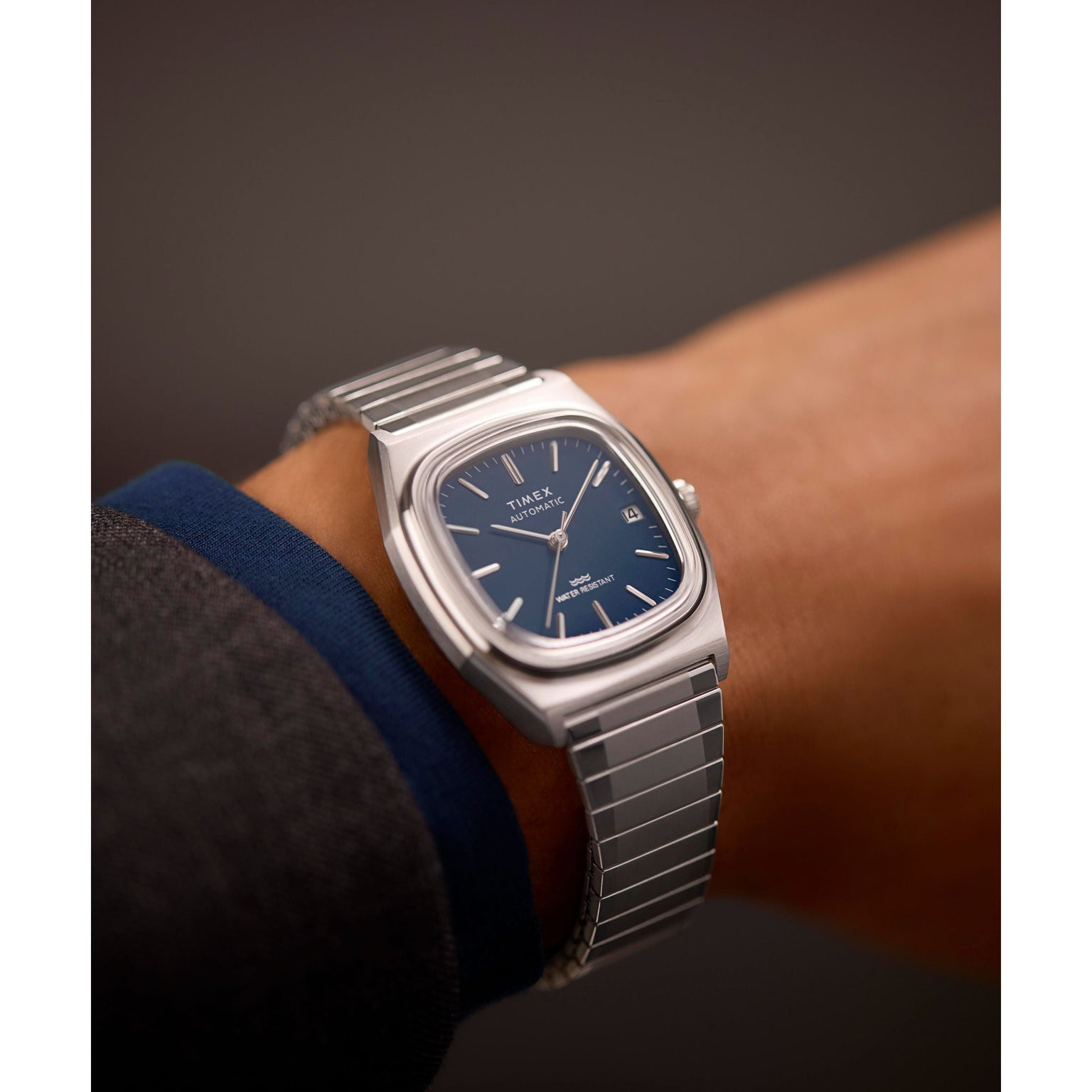 A close-up of a wrist wearing a Timex Automatic 1983 E Line watch with a blue rectangular face, silver hour markers, date window, and stainless-steel expansion band. The person has on a dark sleeve with a blue cuff.