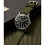 The Timex G SS AU SW MK1 40 BLK DL GRN STP 25, featuring a black dial, white numerals, olive green fabric strap, and stainless steel case, is shown on a textured green surface beside a black box.