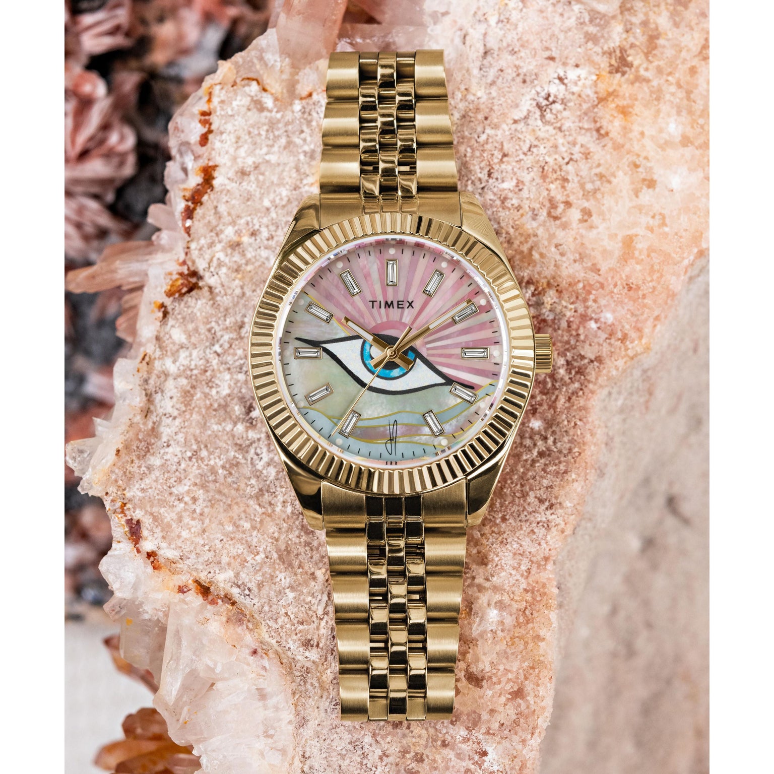A Timex x Jacquie Aiche wristwatch with a gold link band and multicolored mother-of-pearl dial sits on a large crystal formation in pink and beige tones.
