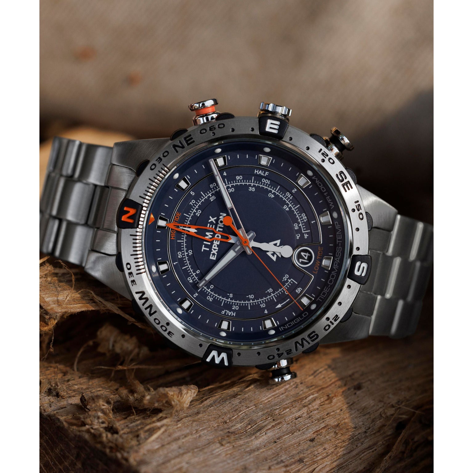The Timex Expedition Tide-Temp-Compass watch, with its black dial, compass bezel, and orange accents, rests on rough wooden planks. Multiple dials and date display add to its rugged yet refined appeal.