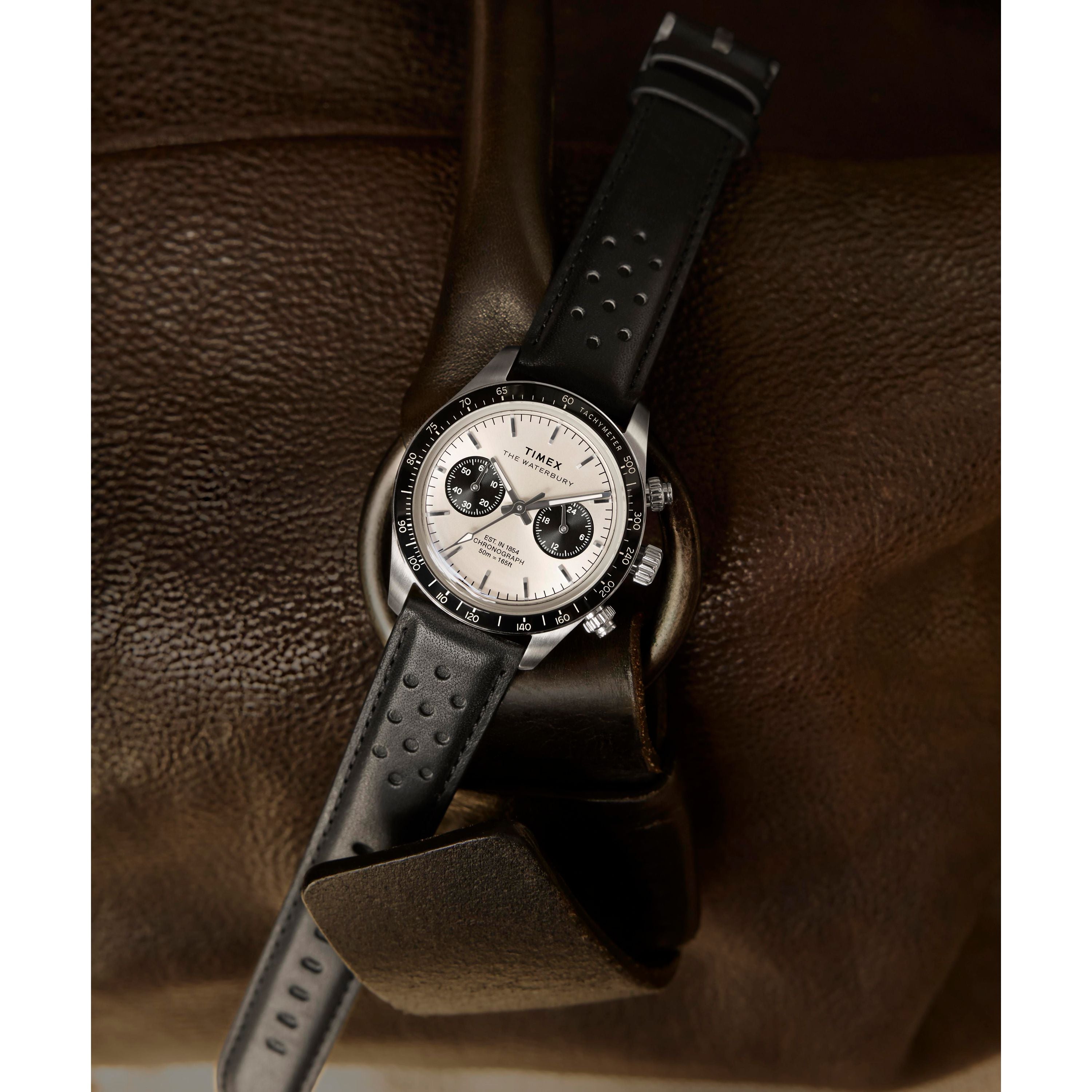 The Timex Waterbury Heritage Chronograph, a silver and black watch with a leather band, rests on brown leather. Its face shows three subdials, a tachymeter bezel, and the time set to about 10:09.