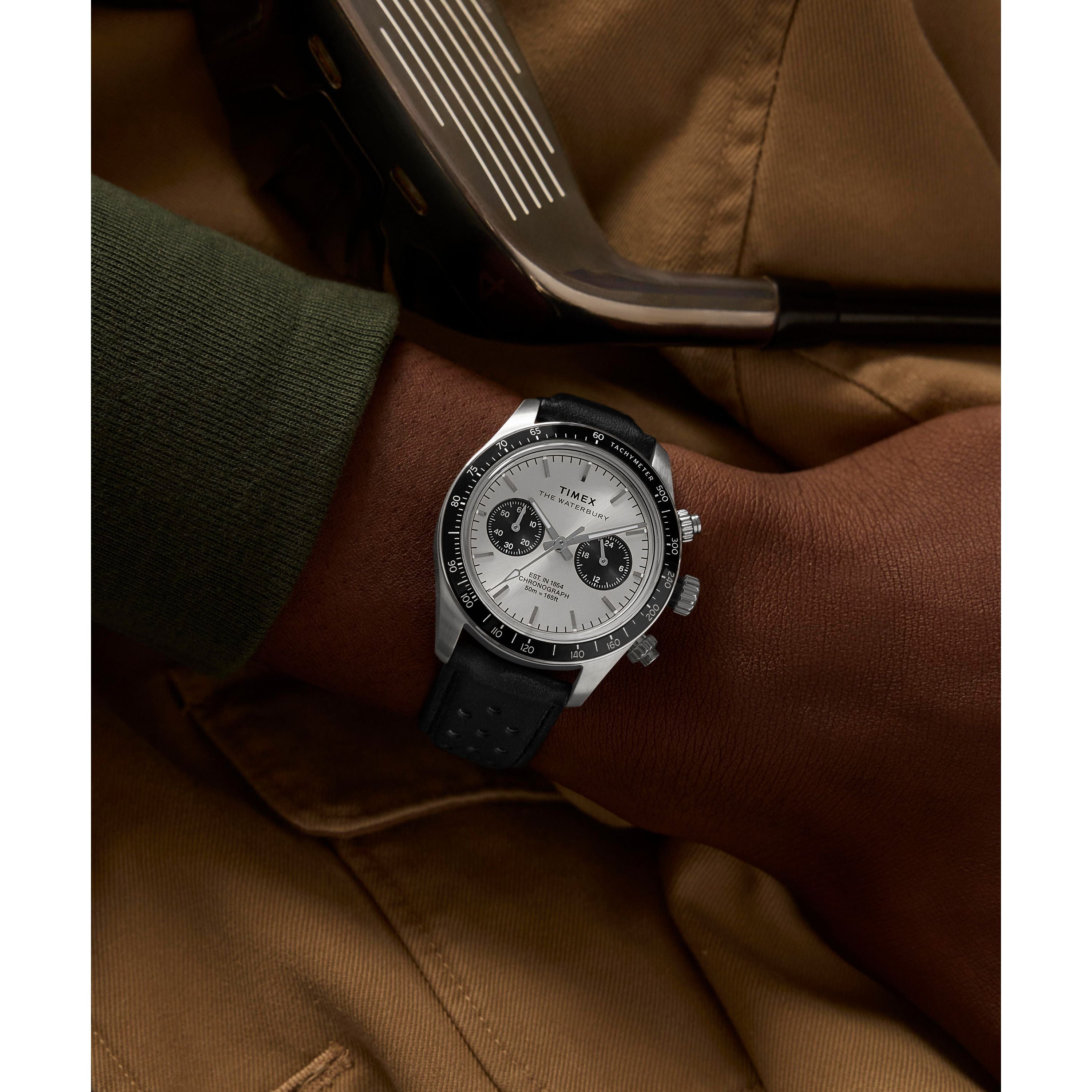 Wearing a Timex Waterbury Heritage Chronograph with a black strap and tachymeter bezel, someone rests their arm on tan fabric while holding a golf club.
