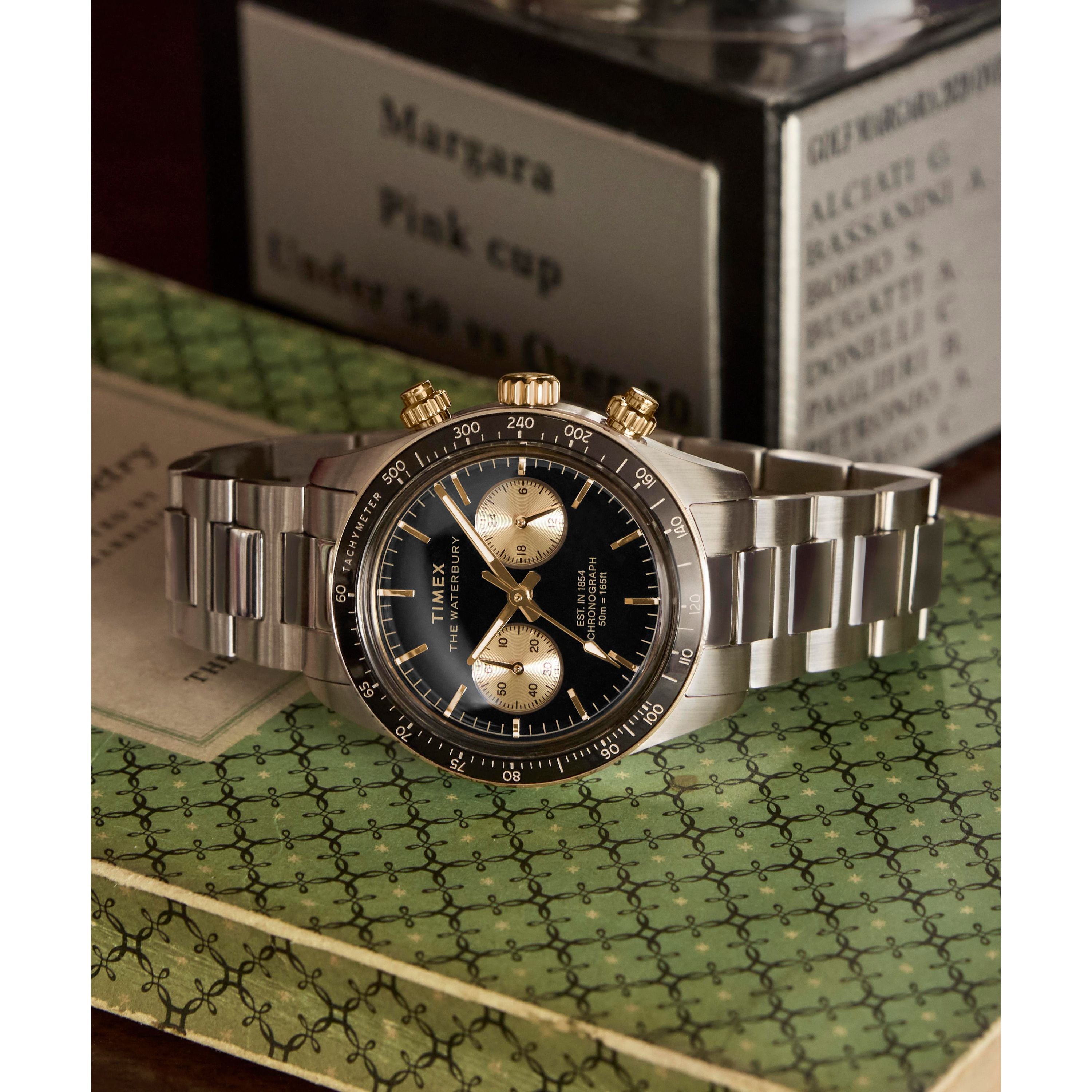 The Timex Waterbury Heritage Chronograph, featuring a silver stainless steel case and black and gold tachymeter bezel with two subdials, is displayed on a green patterned book with blurred books and signs in the background.