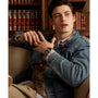 A young man in a denim jacket and a Timex Waterbury Heritage Chronograph sits on a sofa, gazing sideways. Behind him, shelves lined with books evoke a cozy library-like atmosphere.
