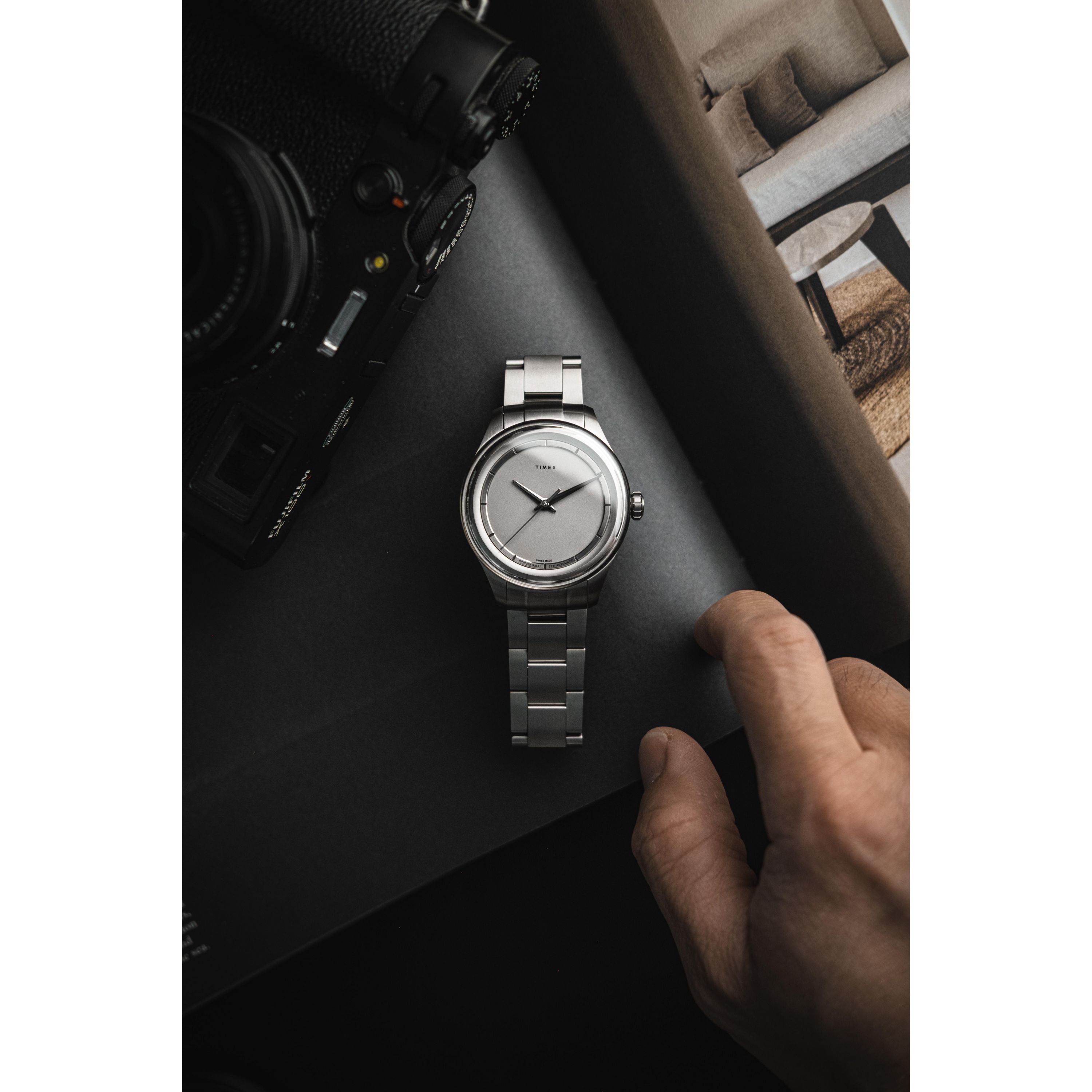 A Timex Giorgio Galli S2Ti Automatic, 38mm titanium with a silver metal band, rests on a dark surface next to a camera and book as a hand reaches for it.