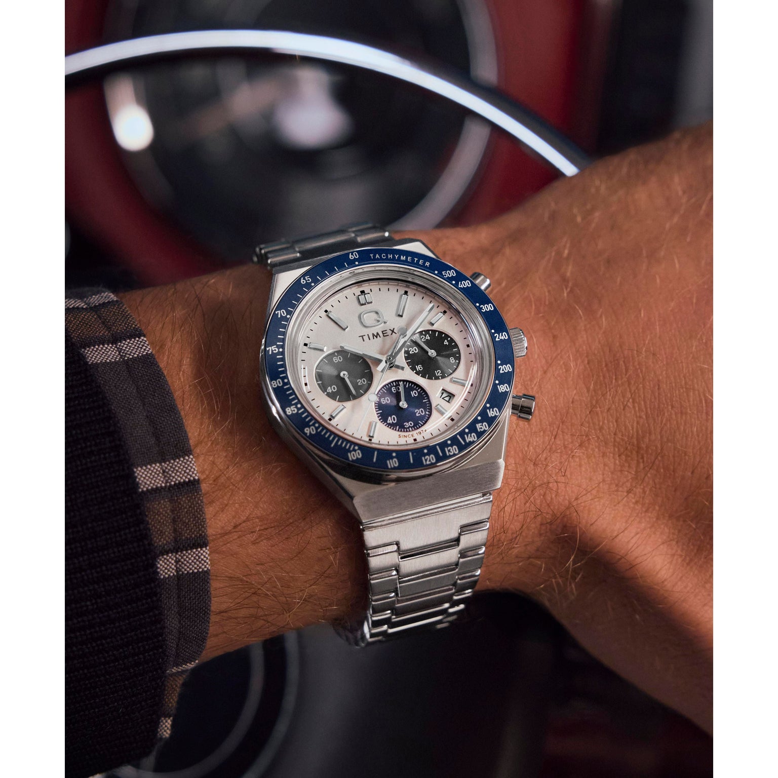 A close-up shows a person’s wrist inside a car, wearing a Timex Q Chronograph racing watch with a blue bezel, white face, and stainless steel bracelet; a plaid shirt sleeve is partially visible.