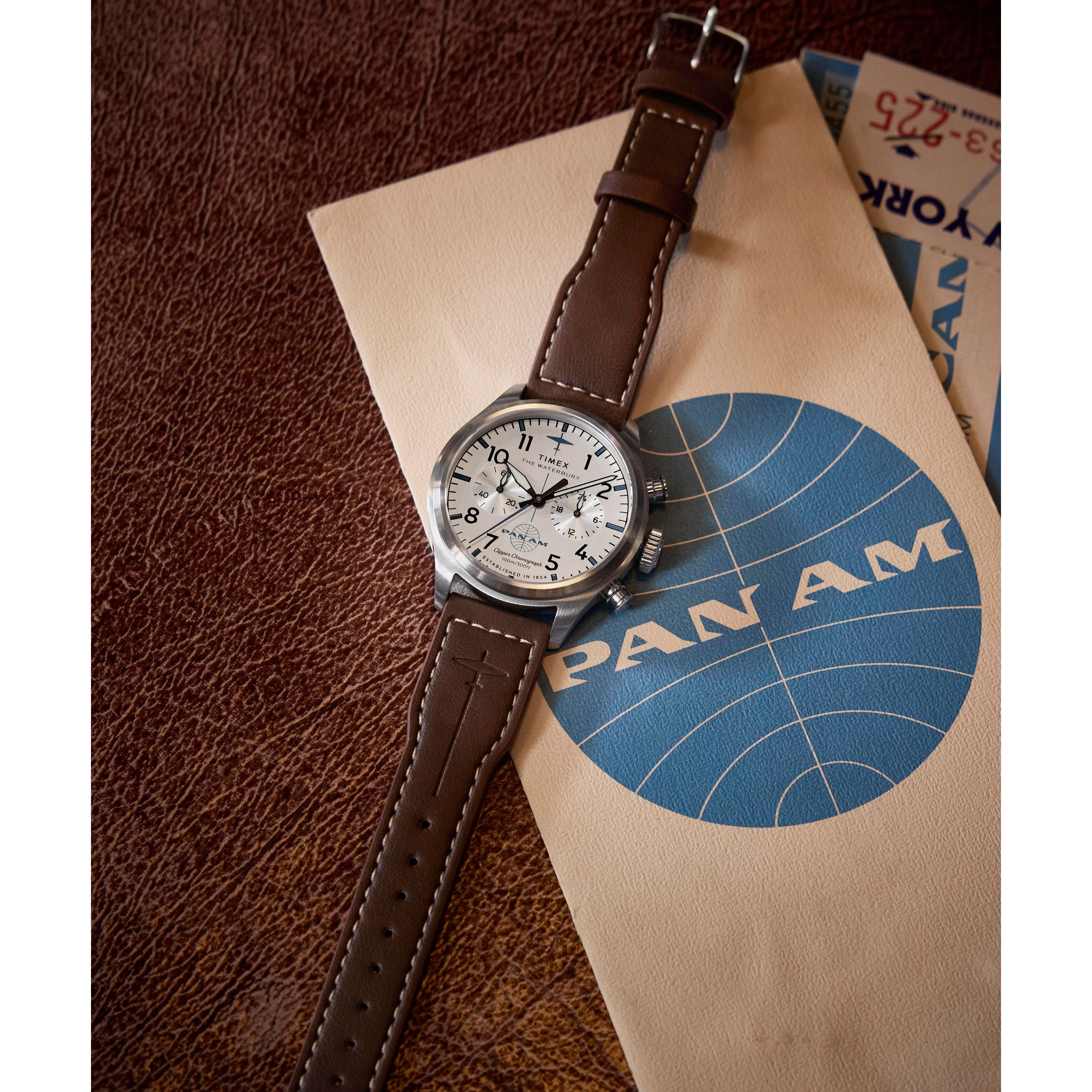 The Timex G SS QZ SW WATERBURY TRAD 41 WHITE DL 25 pilot watch, featuring a brown leather strap and stainless-steel case, sits on a vintage Pan Am envelope with part of a retro airline ticket peeking out.
